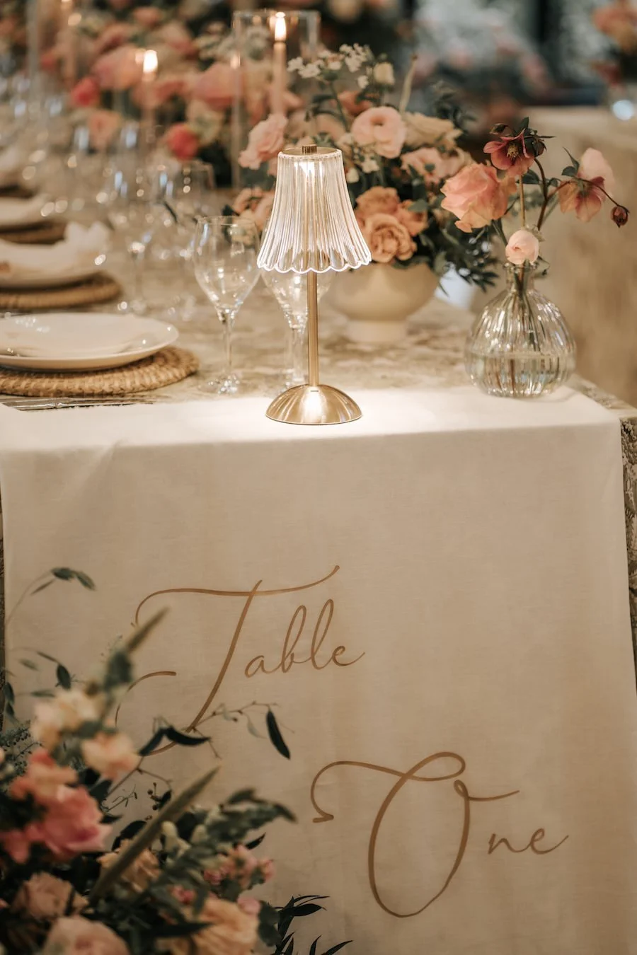 Elegant wedding reception table with pink and cream flowers, a small brass lamp, and table setting. A cream tablecloth has 'Table One' written in gold script.
