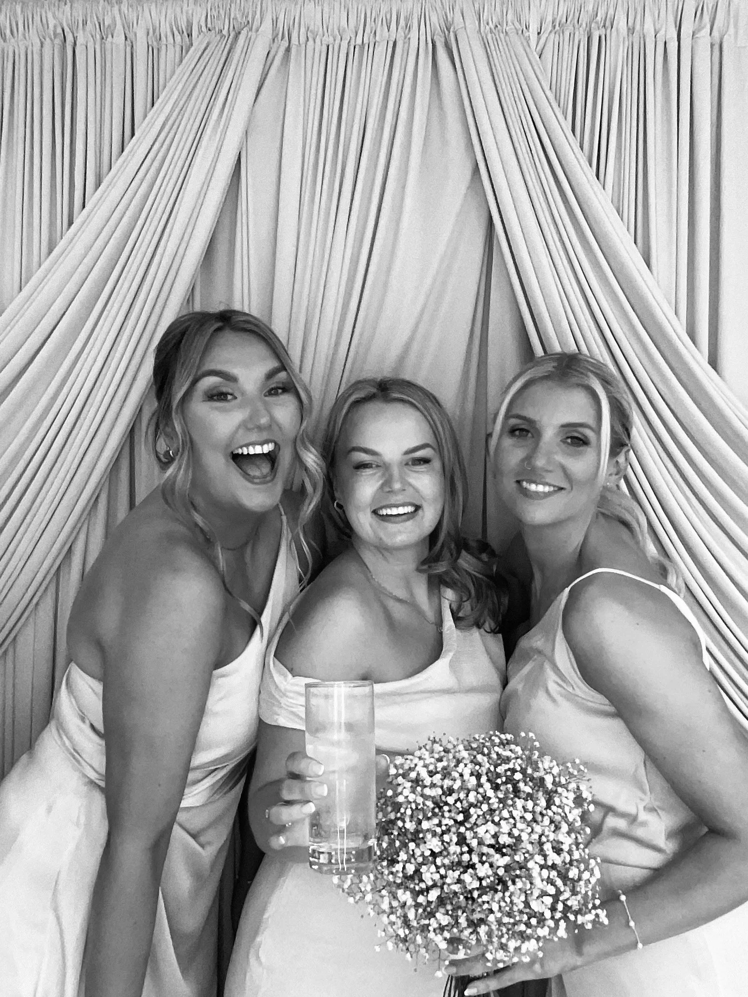 Three women in dresses celebrating at a party, with one woman holding a glass and a bouquet of balloons on the table.