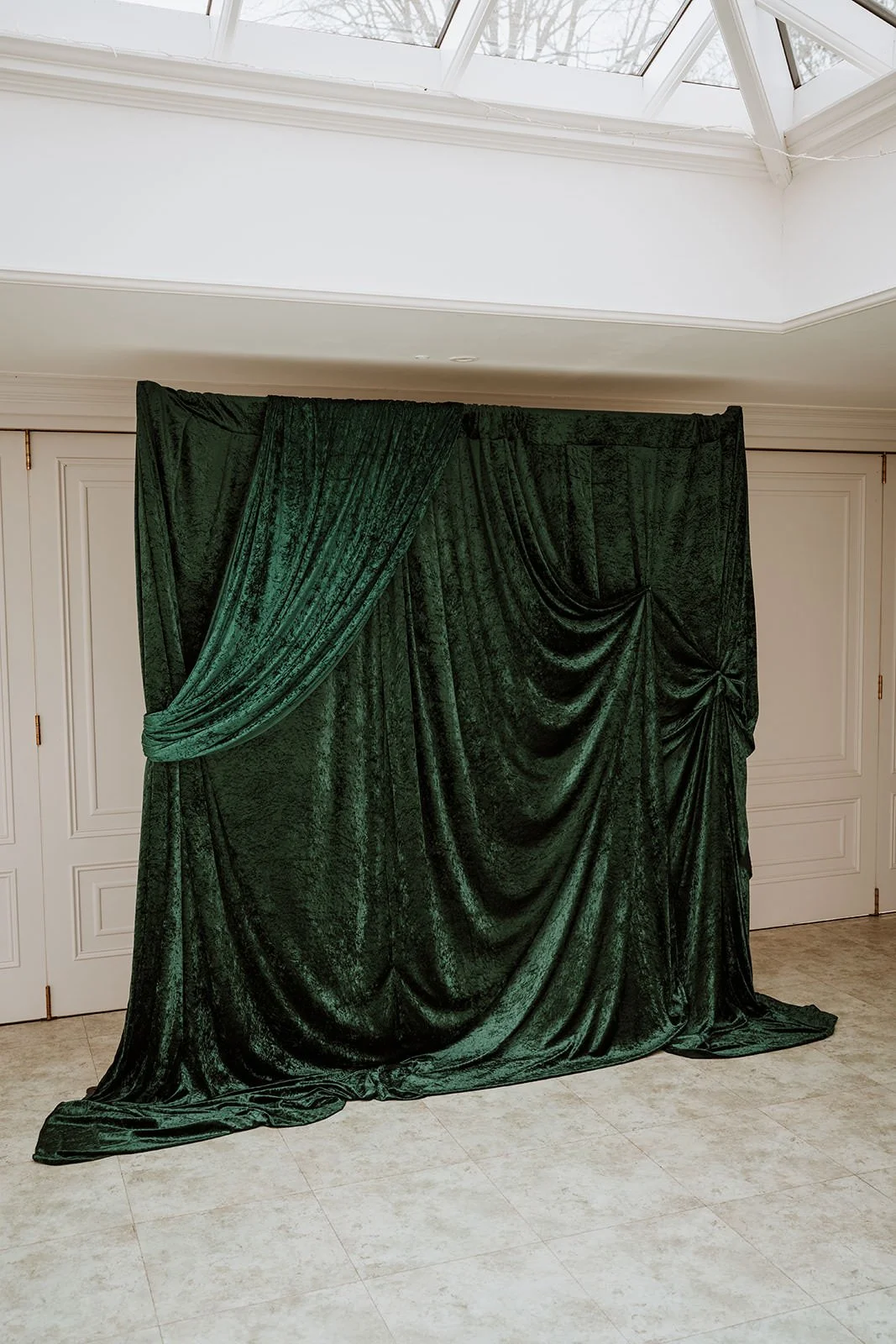 A dark green velvet curtain draped and gathered, hanging in front of white paneled doors inside a room with a tile floor and a skylight ceiling.