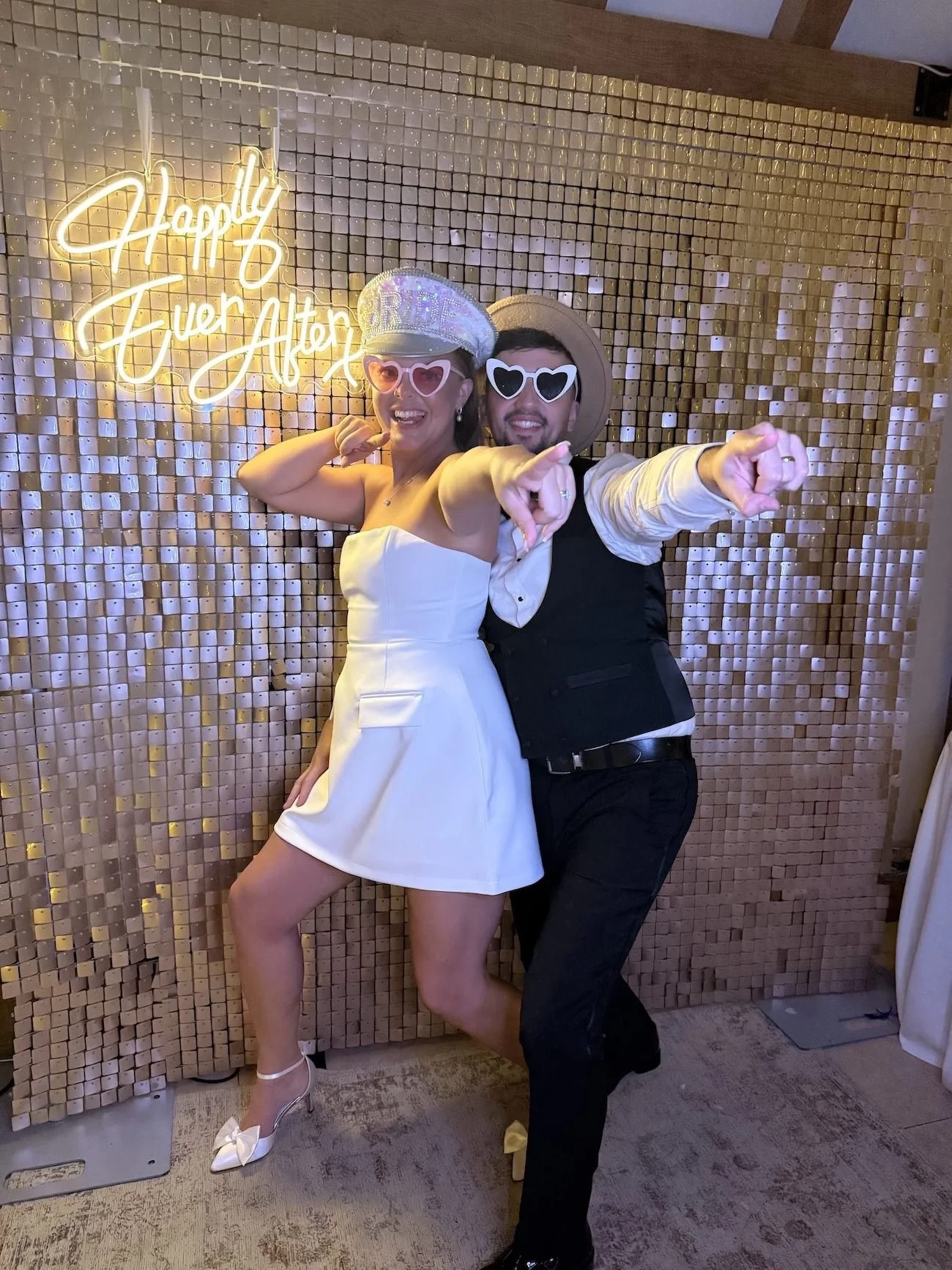 A happy woman in a white dress and heels, and a man in a black vest, dress shirt, and hat, both wearing heart-shaped sunglasses, celebrating at a party with a 
