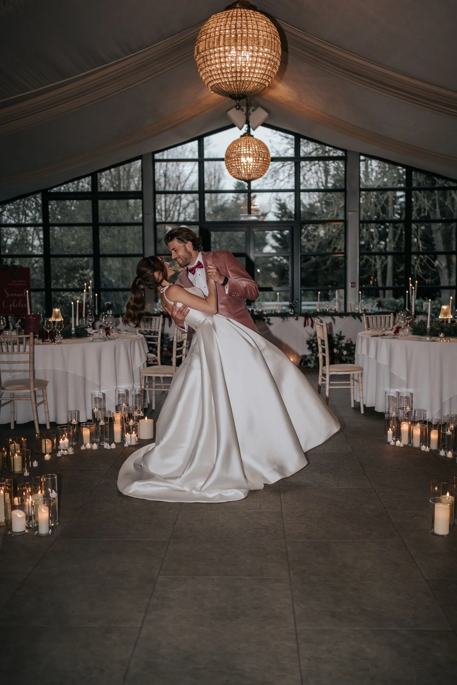 A bride and groom dancing at a wedding reception in a decorated venue with candles on the floor, tables with candles and wine glasses, large windows showing trees outside, and warm lighting from hanging lamps.