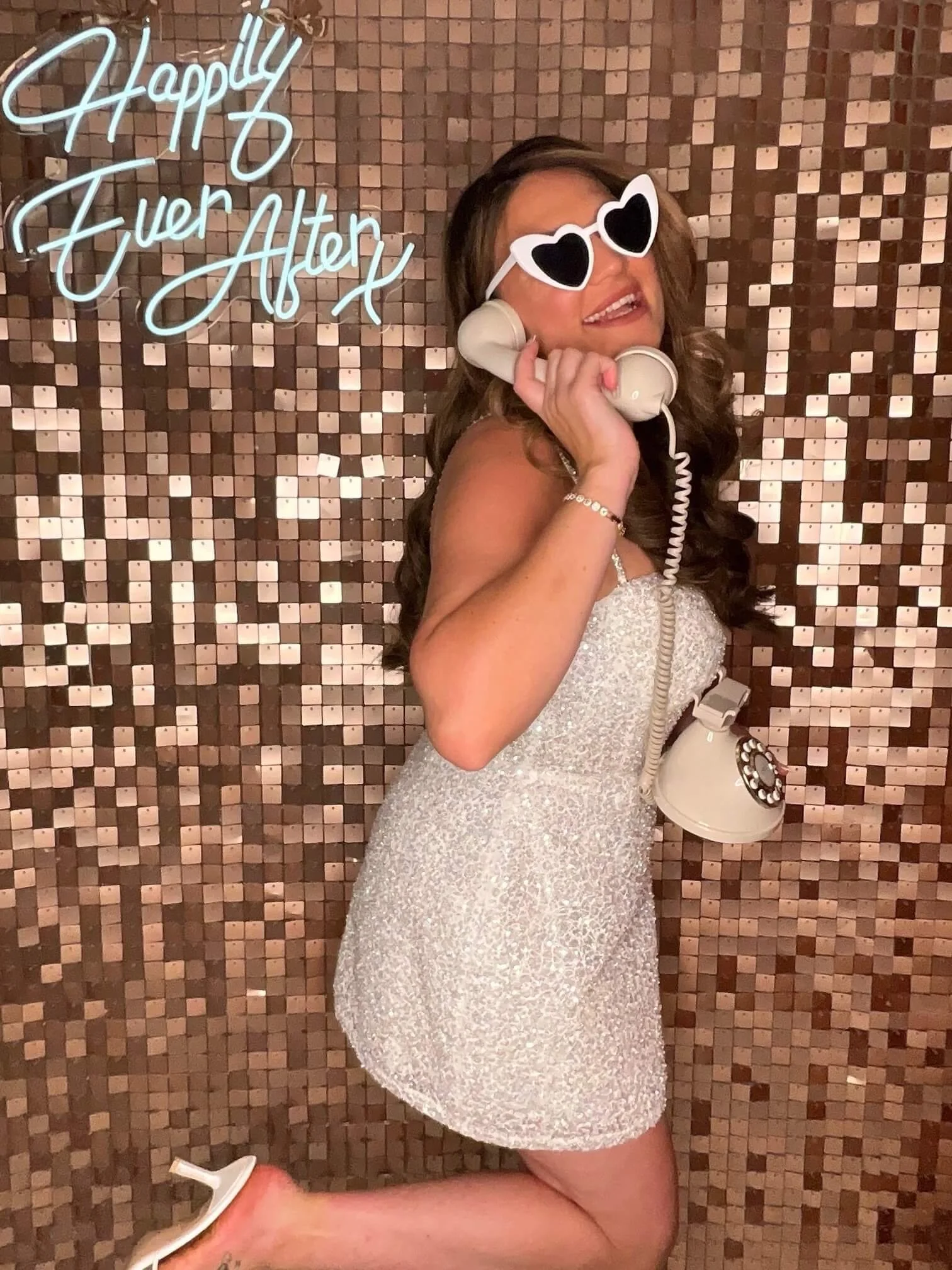 Woman in a sparkly white dress with heart-shaped sunglasses, holding a beige telephone, standing against a mosaic tile wall with a neon sign that reads 'HAPPY Ever After'.