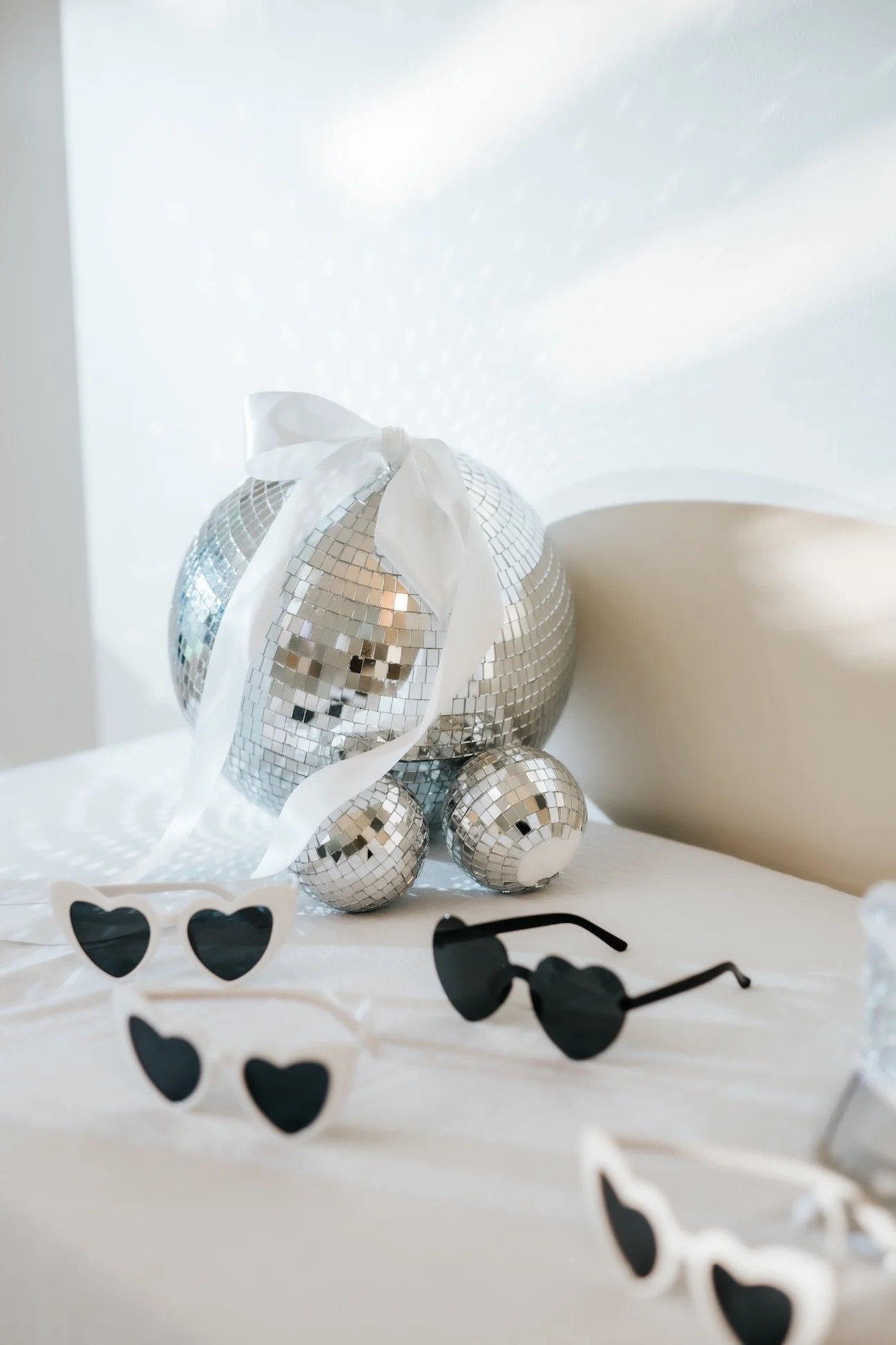 A disco ball shaped decoration with a white ribbon on top, surrounded by several smaller disco ball decorations of different sizes, and a pair of white sunglasses with black heart-shaped lenses and black sunglasses on a white surface.
