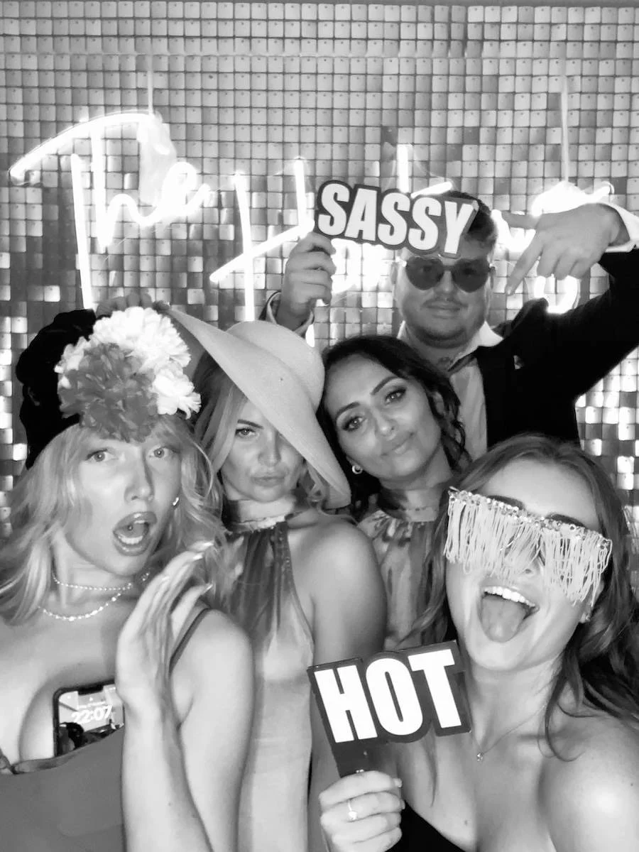 Group of five young adults at a party, wearing fun accessories and holding signs that read 'SASSY' and 'HOT', posing in front of a neon light backdrop.
