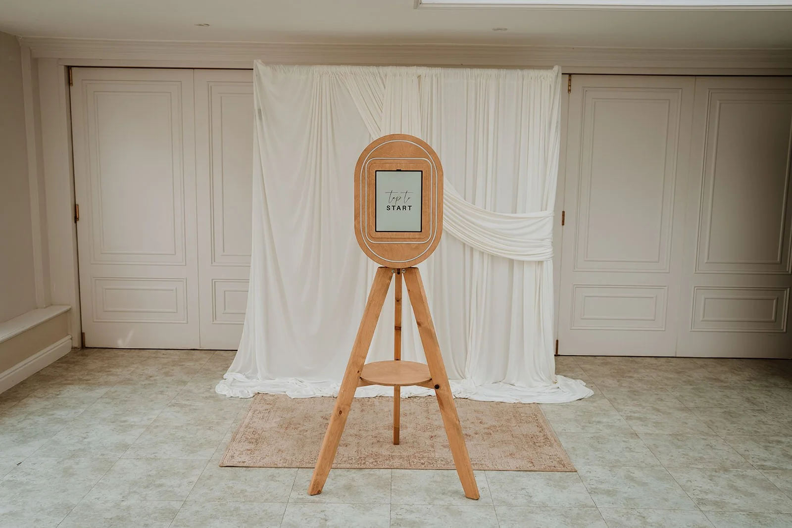 A wooden photo booth frame on a tripod stand with an electronic screen displaying 'Tap to Start' in a room with white curtains and paneled white closet doors.