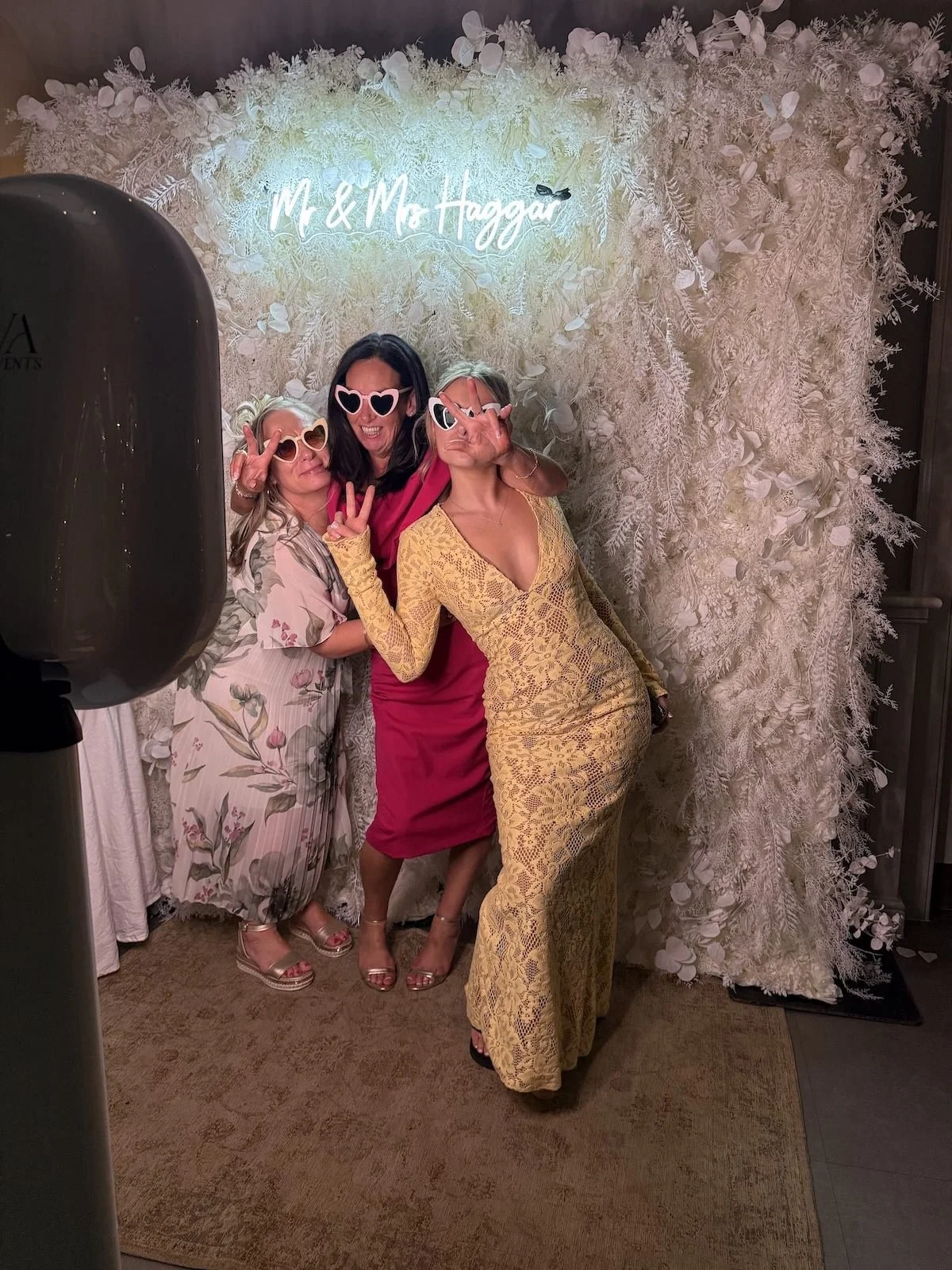 Three women wearing heart-shaped sunglasses, posing playfully in front of a white floral backdrop with a neon sign that reads 'Mr. & Mrs. Haggard.'