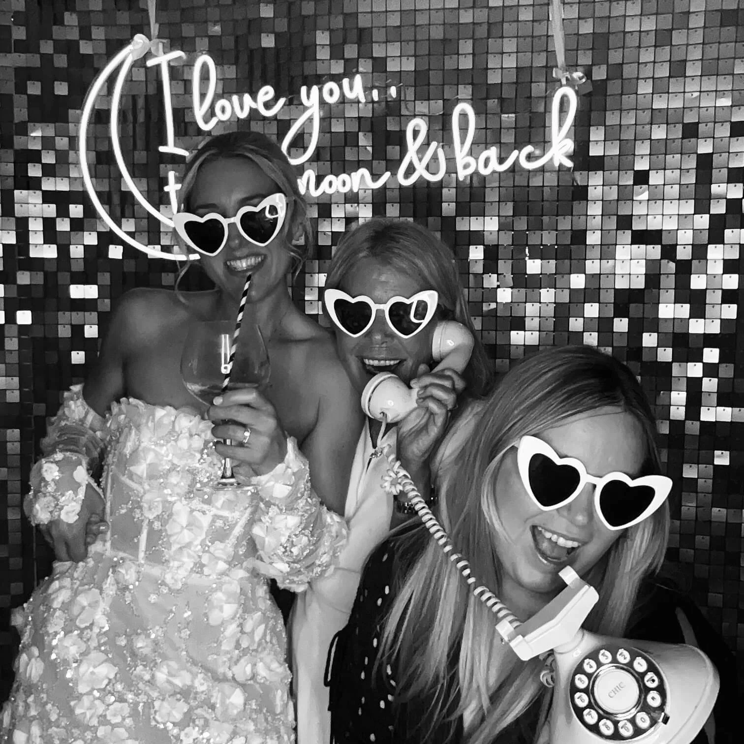 Three women at a party wearing heart-shaped sunglasses, holding phones and a drink, posing in front of a decorative wall with a neon sign that says 'I love you, moon & back'.