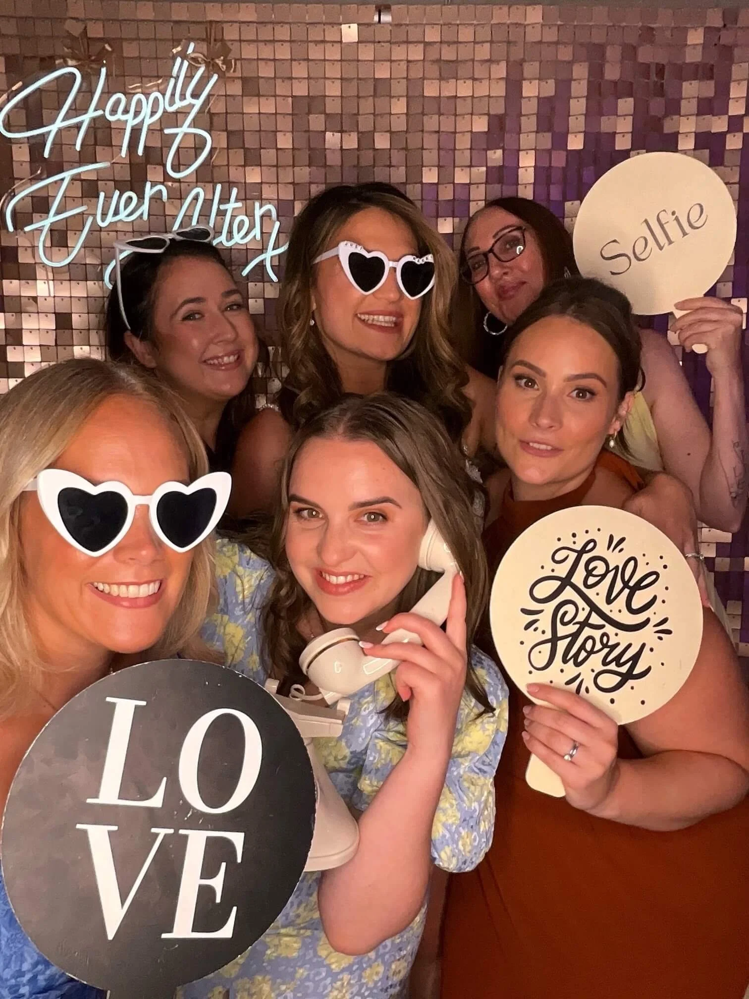 A group of women at a celebration, holding various signs including 'Happily Ever After,' 'Selfie,' and 'Love Story.' They are smiling and wearing accessories like sunglasses and glasses in front of a shiny, mirrored wall with a neon sign.