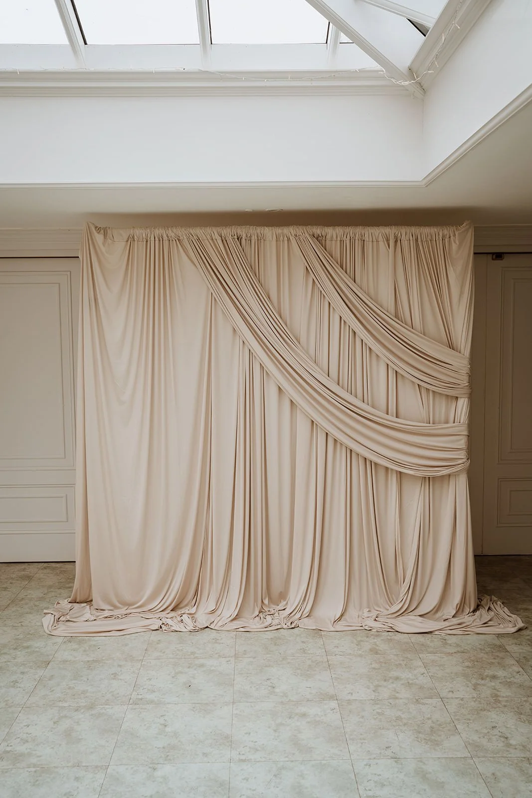 Luxurious beige draped backdrop with elegant folds and twists, covering a window or wall in a formal setting.
