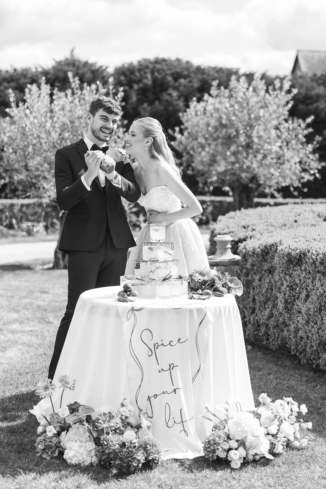 A black-and-white photo of a bride and groom celebrating outdoors. They are pouring champagne into a pyramid of glasses on a table, which is decorated with flowers and a sign that reads 'Spice up your life.' Trees and bushes are in the background.