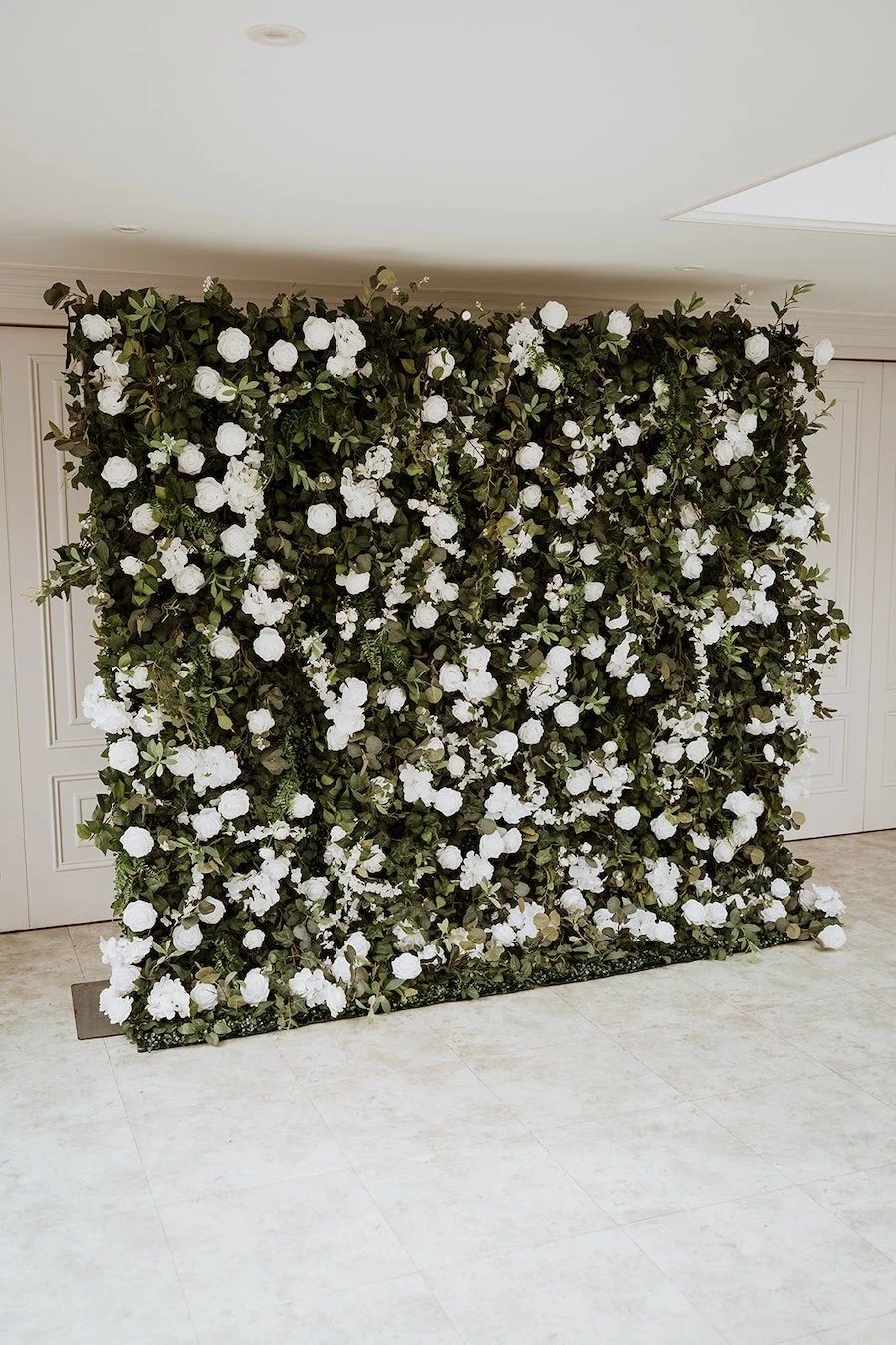 A decorative backdrop covered with white flowers and green foliage, set against a wall with a light-colored floor.