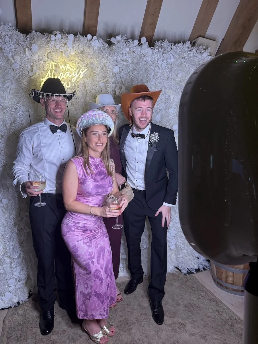 Group of five people celebrating, wearing cowboy hats, with one man in a tuxedo, a woman in a purple dress, and others in semi-formal attire, standing in front of white floral backdrop with illuminated phrase "It Was Always You."