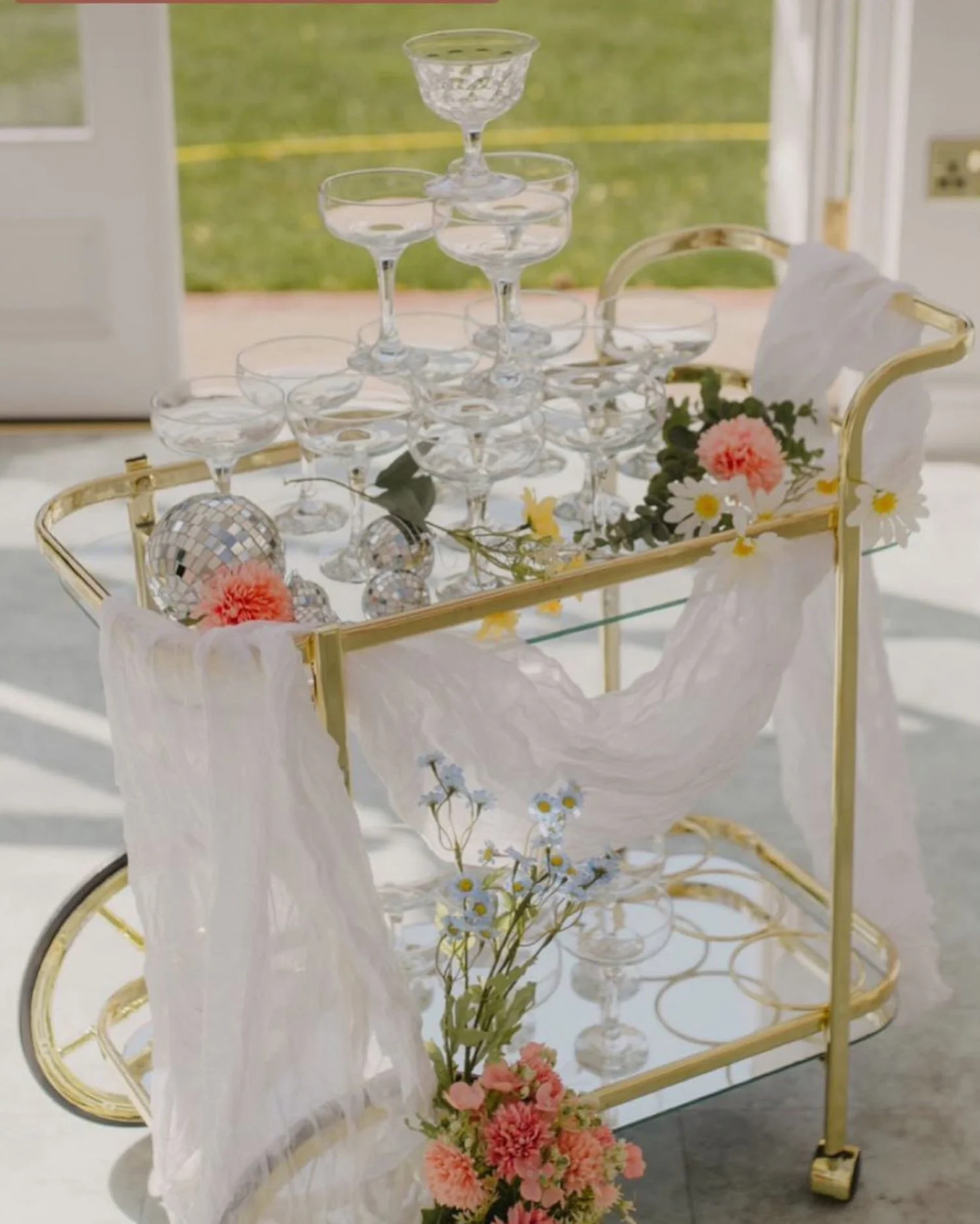 A gold-painted bar cart with a white sheer cloth, decorated with pink carnations, daisies, and baby's breath. On top, there is a champagne tower made of stacked coupe glasses, and disco ball ornaments. Flowers also cascade down from the cart to the f