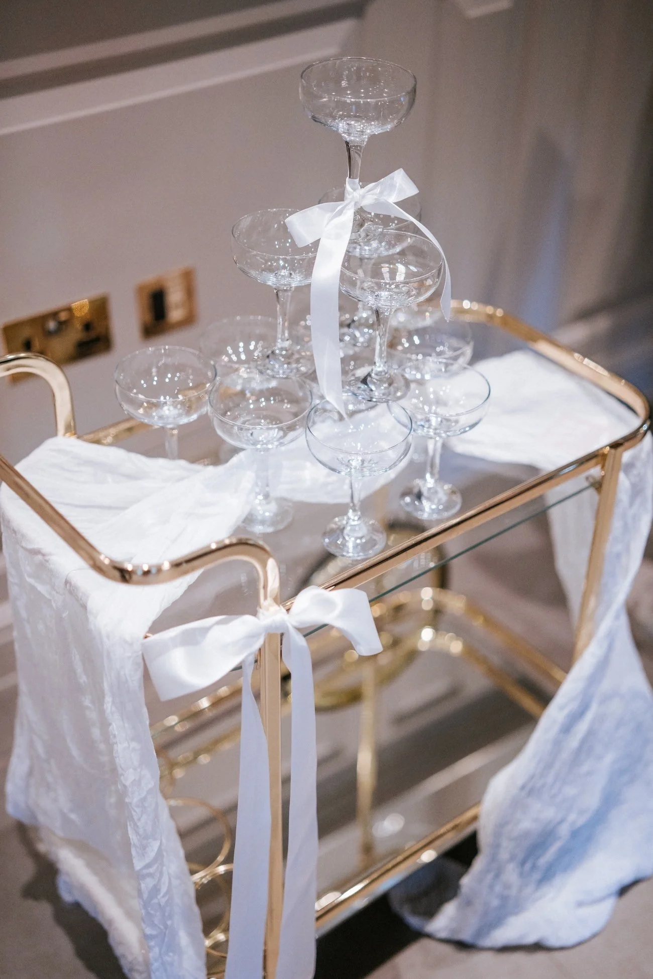 A gold bar cart holding empty coupe glasses stacked in a pyramid with a white ribbon tied around the top glass, draped with white fabric.