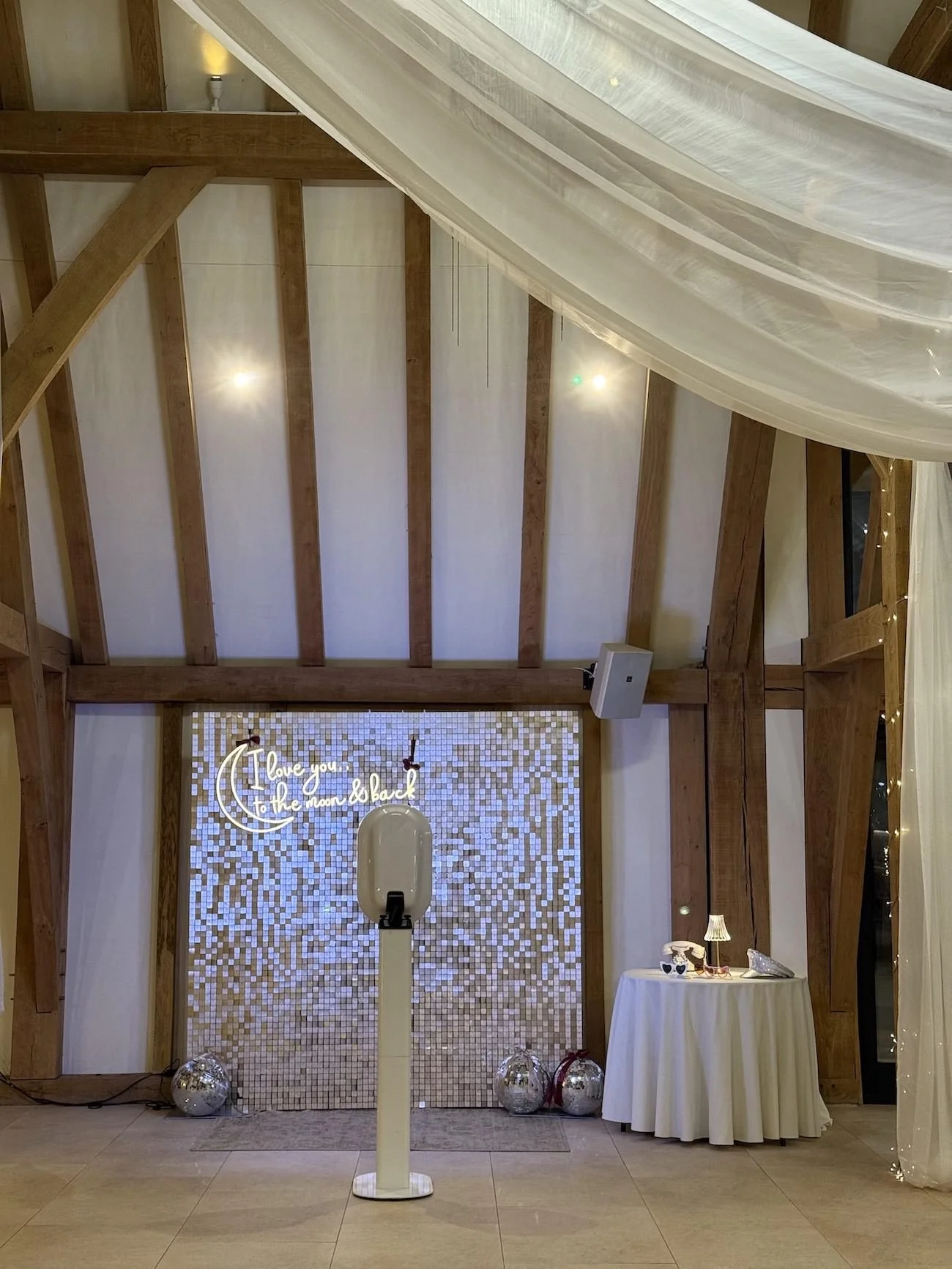 Indoor scene with wooden beams on ceiling, a large white curtain, a lit decorative sign reading 'I love you to the moon & back,' a table with a lamp, decorative items, and balloons on the floor.