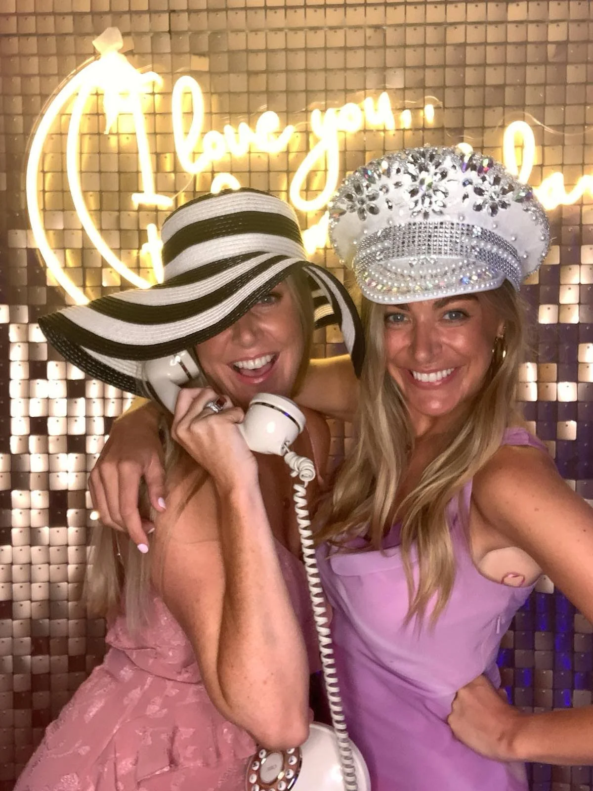 Two women smiling with fancy hats, one with a large black and white striped hat and the other with a jeweled white hat, inside a decorated venue with neon sign in the background.