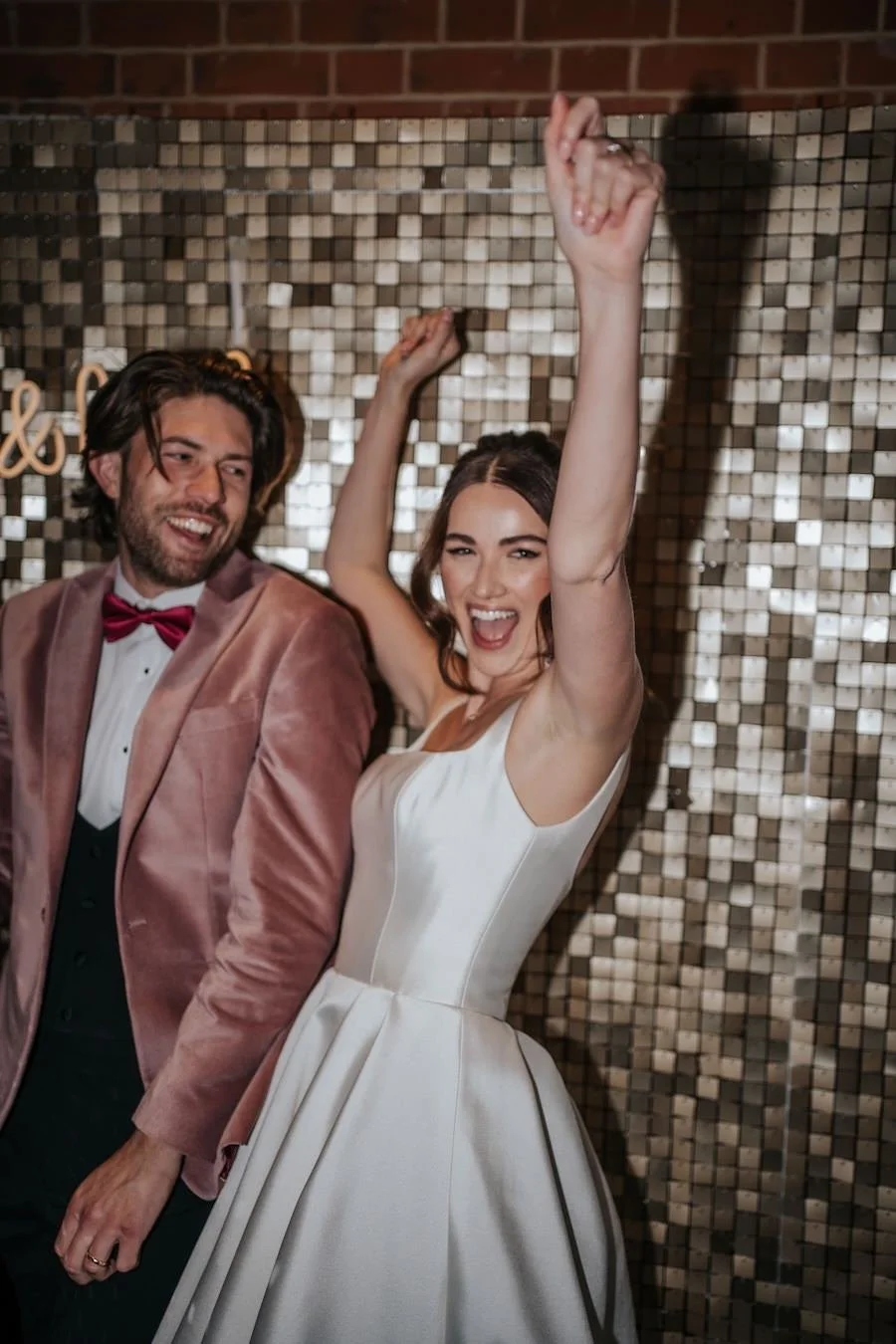 A woman in a white dress celebrating with her arm raised, standing next to a man in a pink velvet blazer and red bow tie, in front of a tiled wall at a festive event.