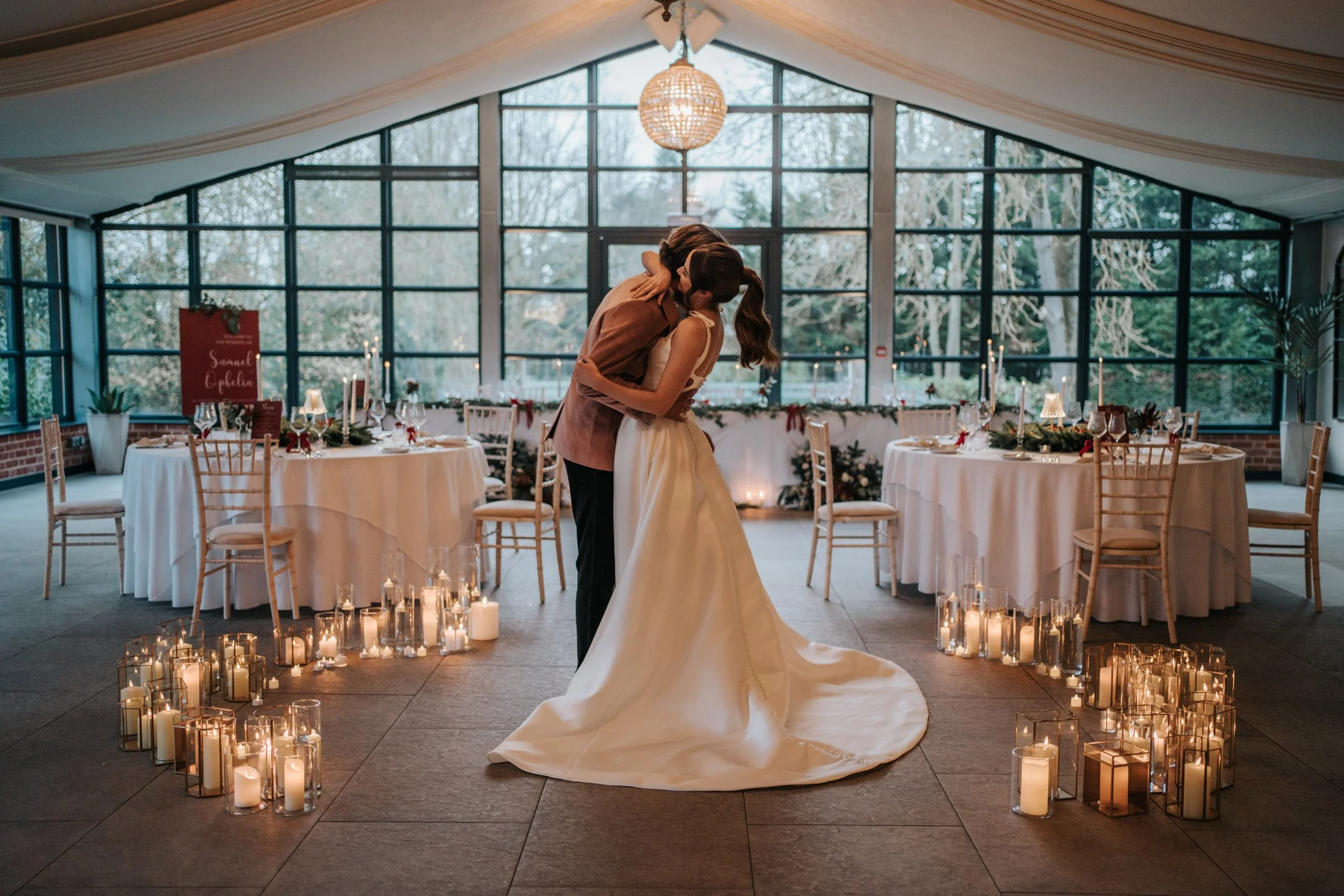 A bride and groom sharing a kiss inside a decorated wedding venue with candles on the floor, round tables, and large windows.