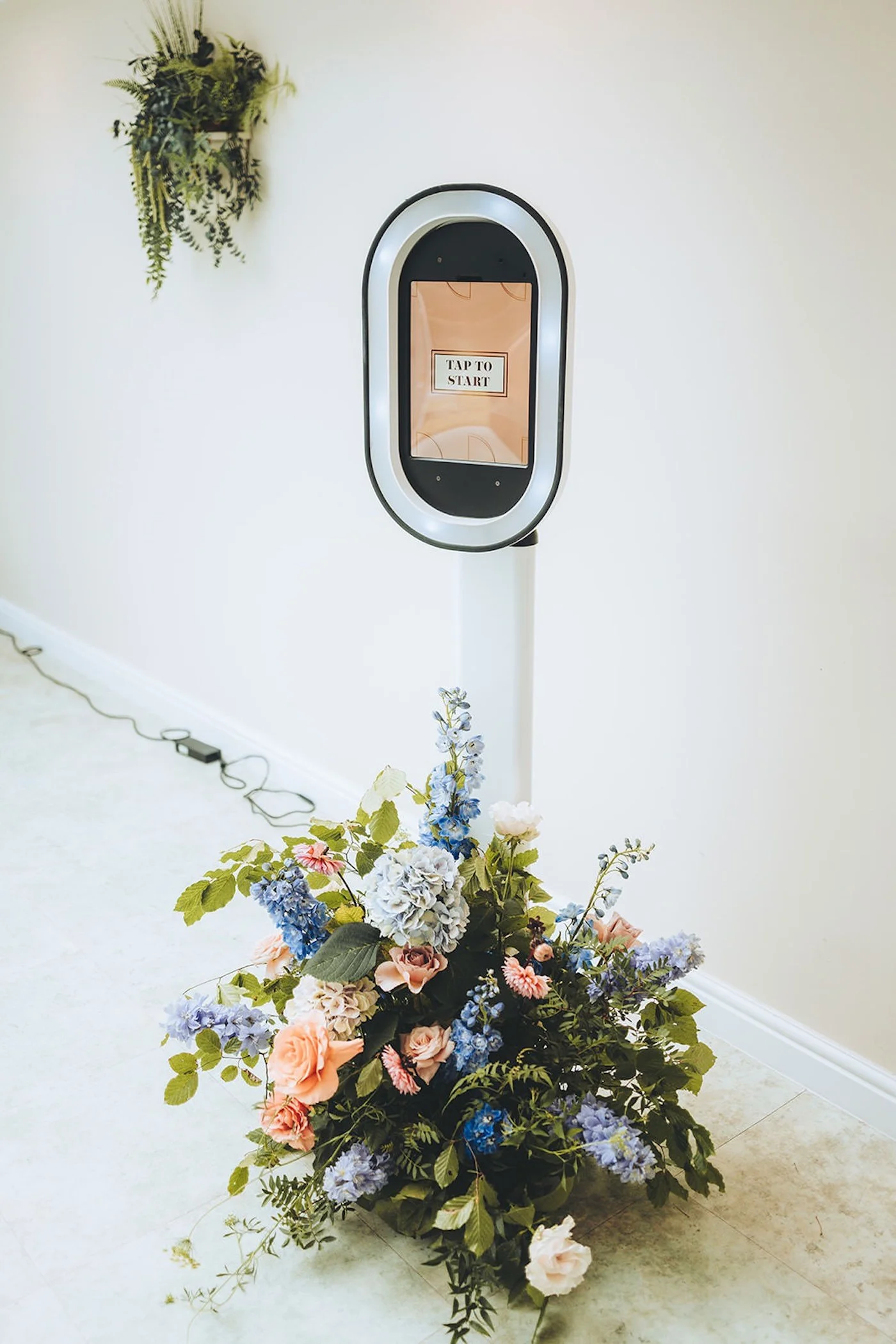 A floral arrangement of various pink, blue, white, and purple flowers with green leaves in front of a white wall. Behind the flowers, there is a black and white oval-shaped device on a stand with a screen displaying a message that says "TAP TO START"