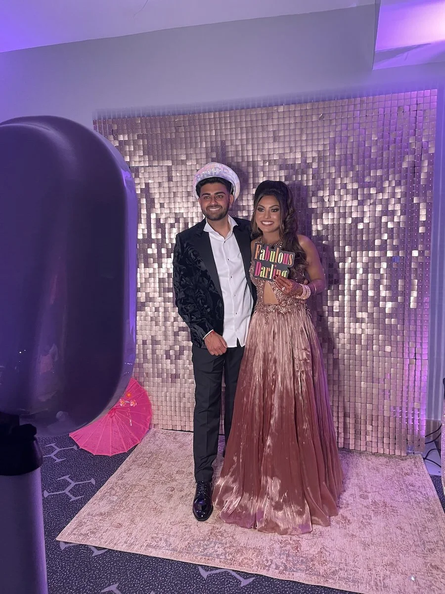 A man and woman dressed in formal attire standing together in front of a decorative backdrop. The woman holds a sign that reads 'Fabulous Darling'.