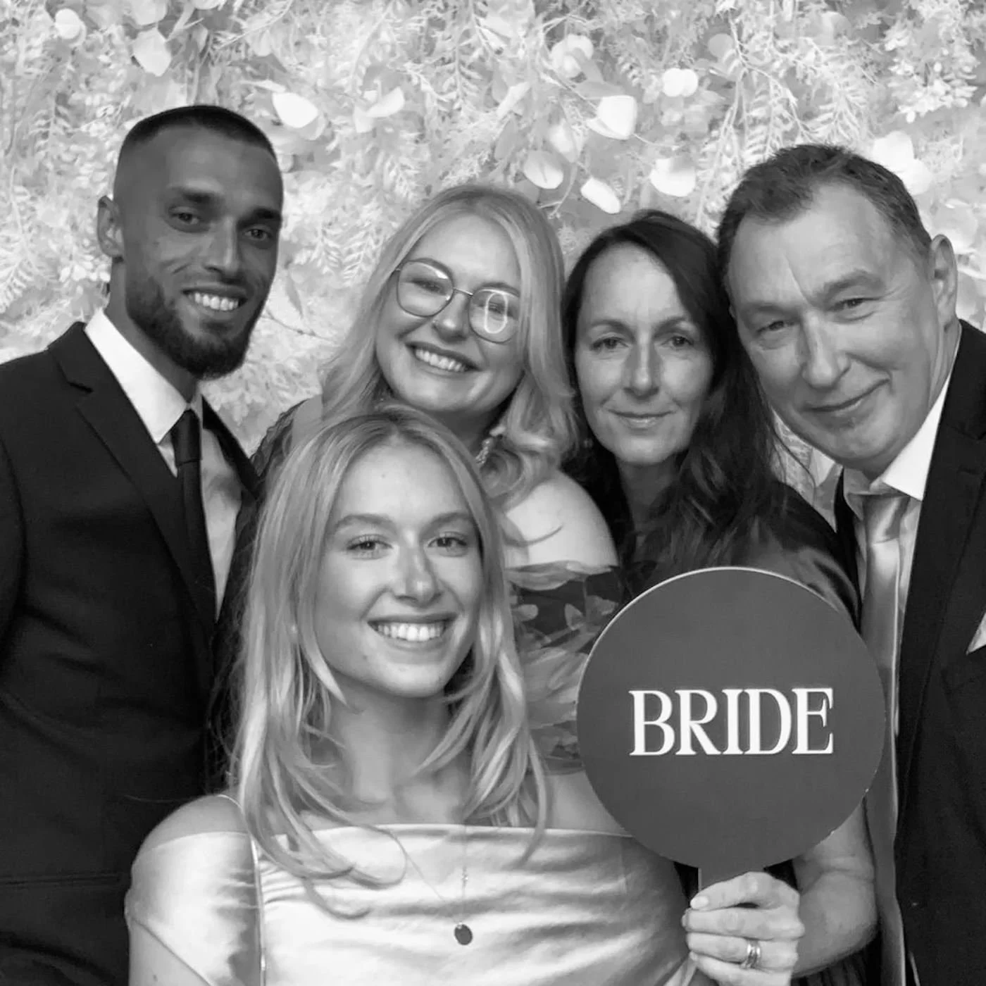 A group of six people, four women and two men, posing for a photo at a wedding or celebration, with one holding a sign that says 'BRIDE'.