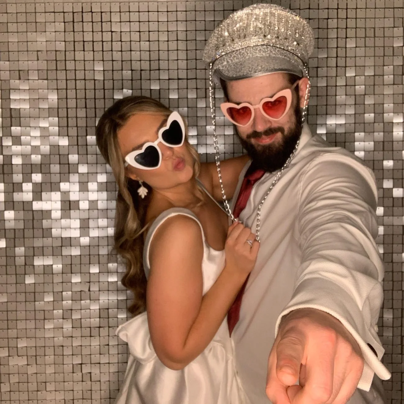 A young couple in party attire taking a selfie in front of a reflective, mosaic tile background. They are wearing large, heart-shaped sunglasses and smiling playfully, with the man pointing at the camera.