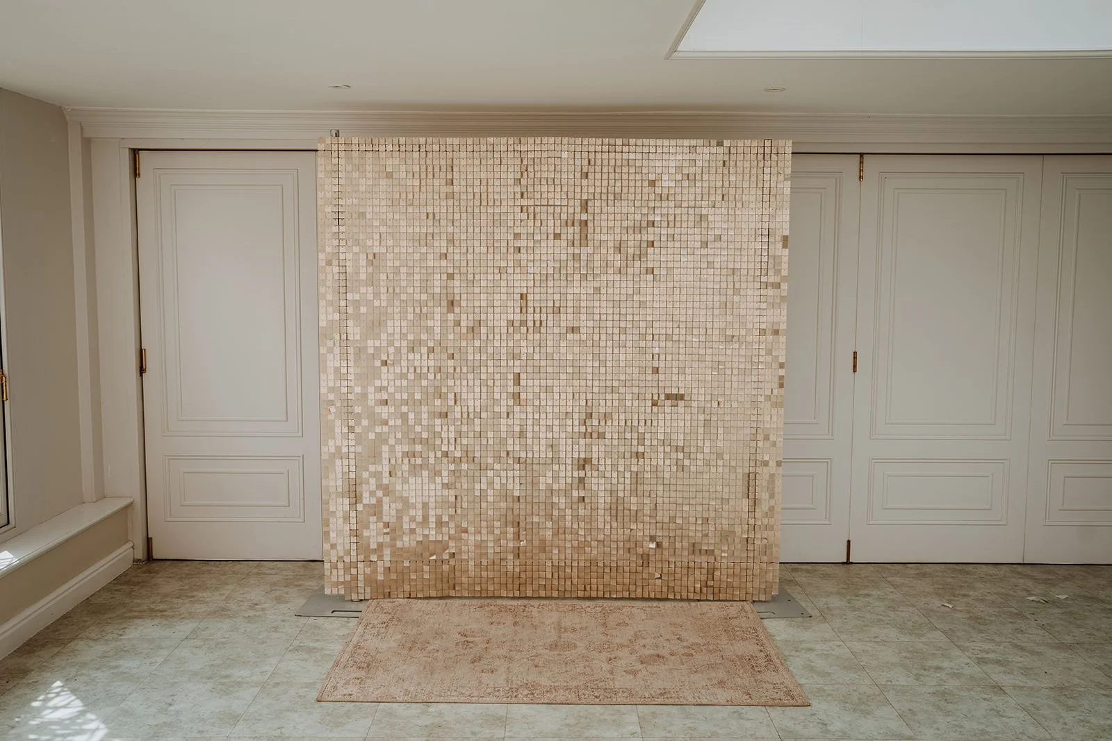 A beige shimmer wall backdrop in front of white paneled closet doors with a beige rug on a tiled floor.