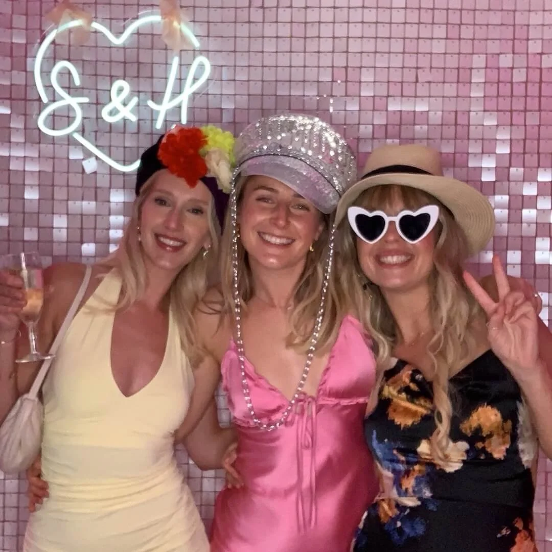 Three women at a celebration, wearing colorful hats and sunglasses, smiling, with a pink mosaic background and a neon sign that reads 'S&H' inside a heart.