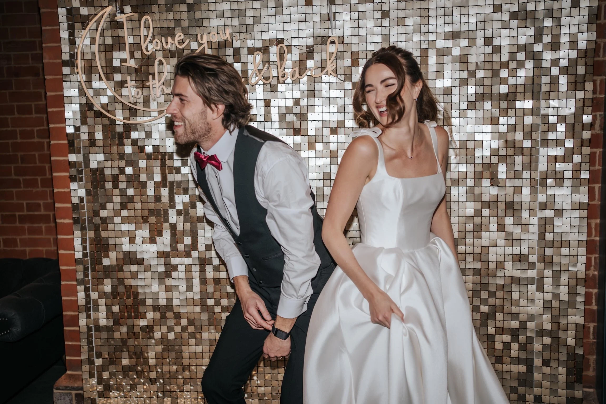 A smiling man in a formal vest, shirt, and bowtie, and a woman in a white dress, are holding hands and leaning back, standing in front of a shiny mosaic wall with a neon sign that reads 'love you to the moon & back.'