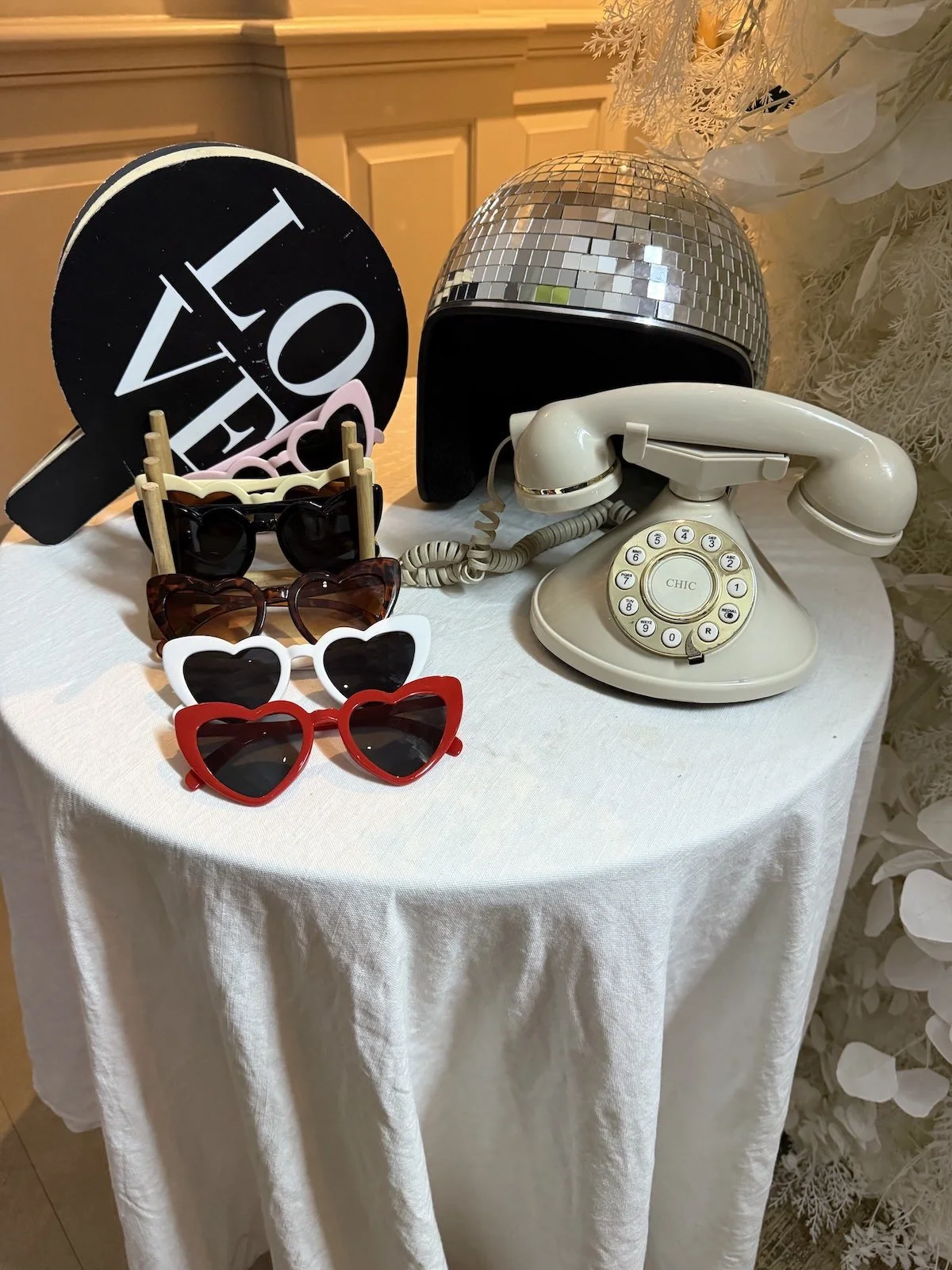 A vintage rotary telephone, a disco ball, a black decorative sign that says 'LOVE', several pairs of sunglasses with heart-shaped and round frames, and white decorative foliage on a white tablecloth-covered table.