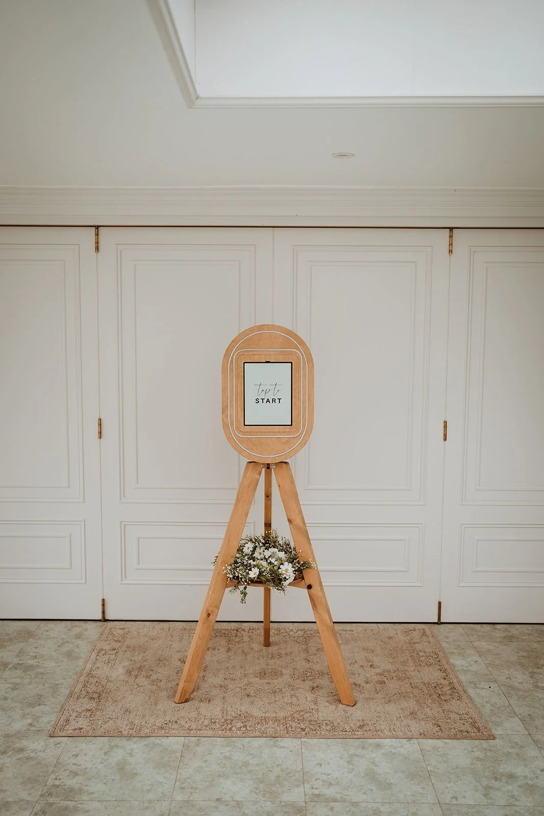 A Vintage style phot booth  a digital screen displaying 'Text START' in black text, a small bouquet of white daisies and greenery at the base, set against white paneled double doors.