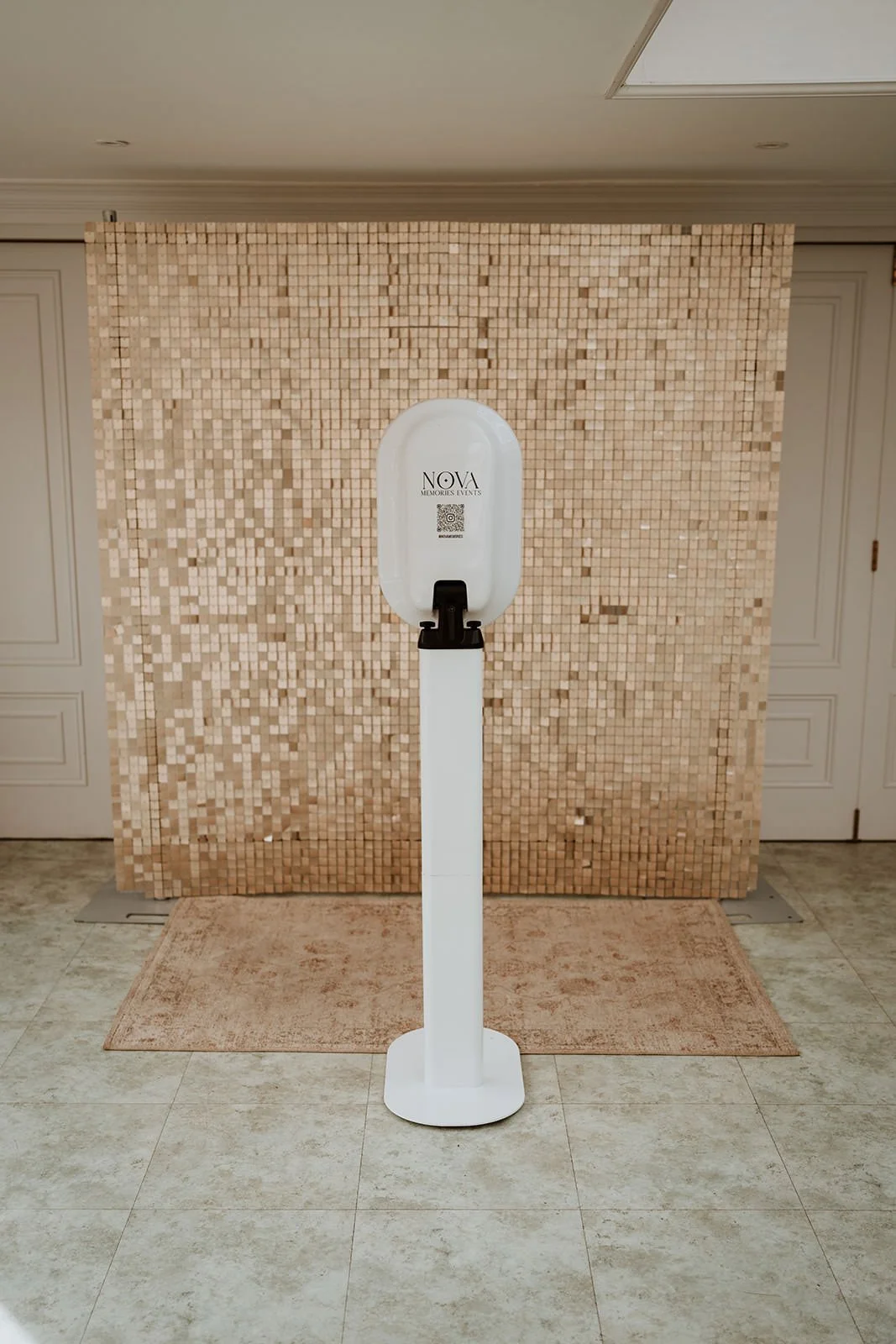 A photo booth in front of a beige mosaic tile wall, with a small pink rug on a tiled floor beneath it.