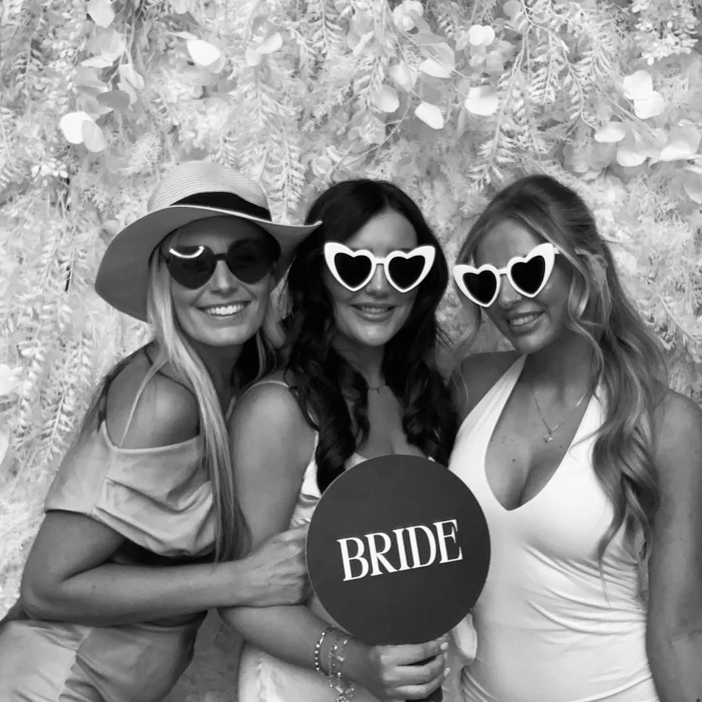Three women at a wedding celebration wearing sunglasses, with the bride holding a sign that says 'BRIDE' and a round sign, posing in front of a floral backdrop.