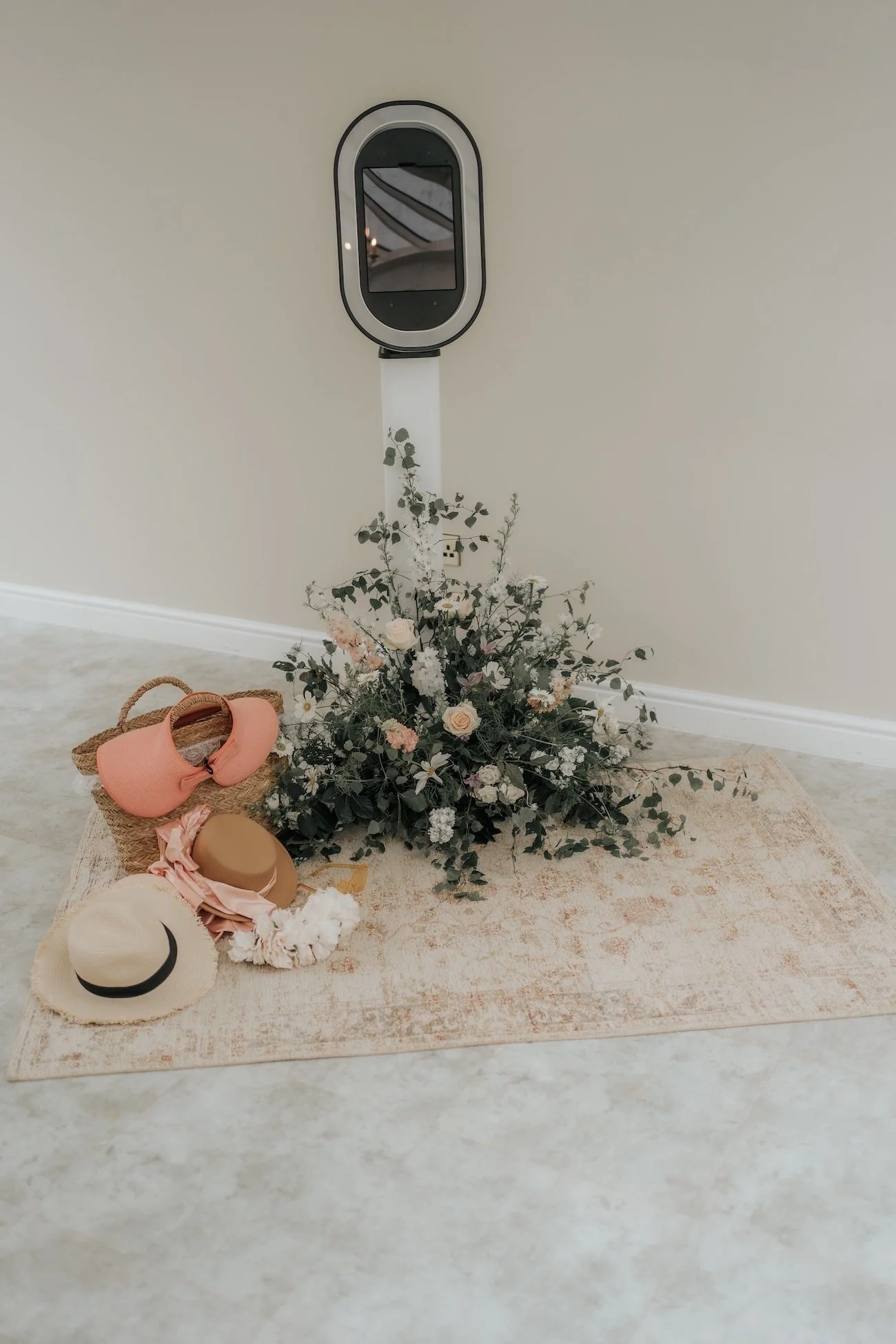 A beige rug with a bouquet of flowers and hats placed on it, set against a plain wall with a face mask hanging in the background.