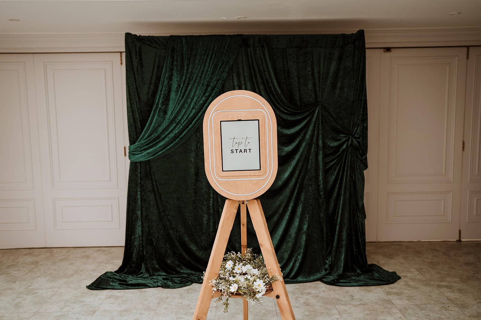 Photo of a photo booth backdrop with dark green velvet curtains, a wooden easel with a sign that says 'tap to START', and a small bouquet of white flowers at the base.