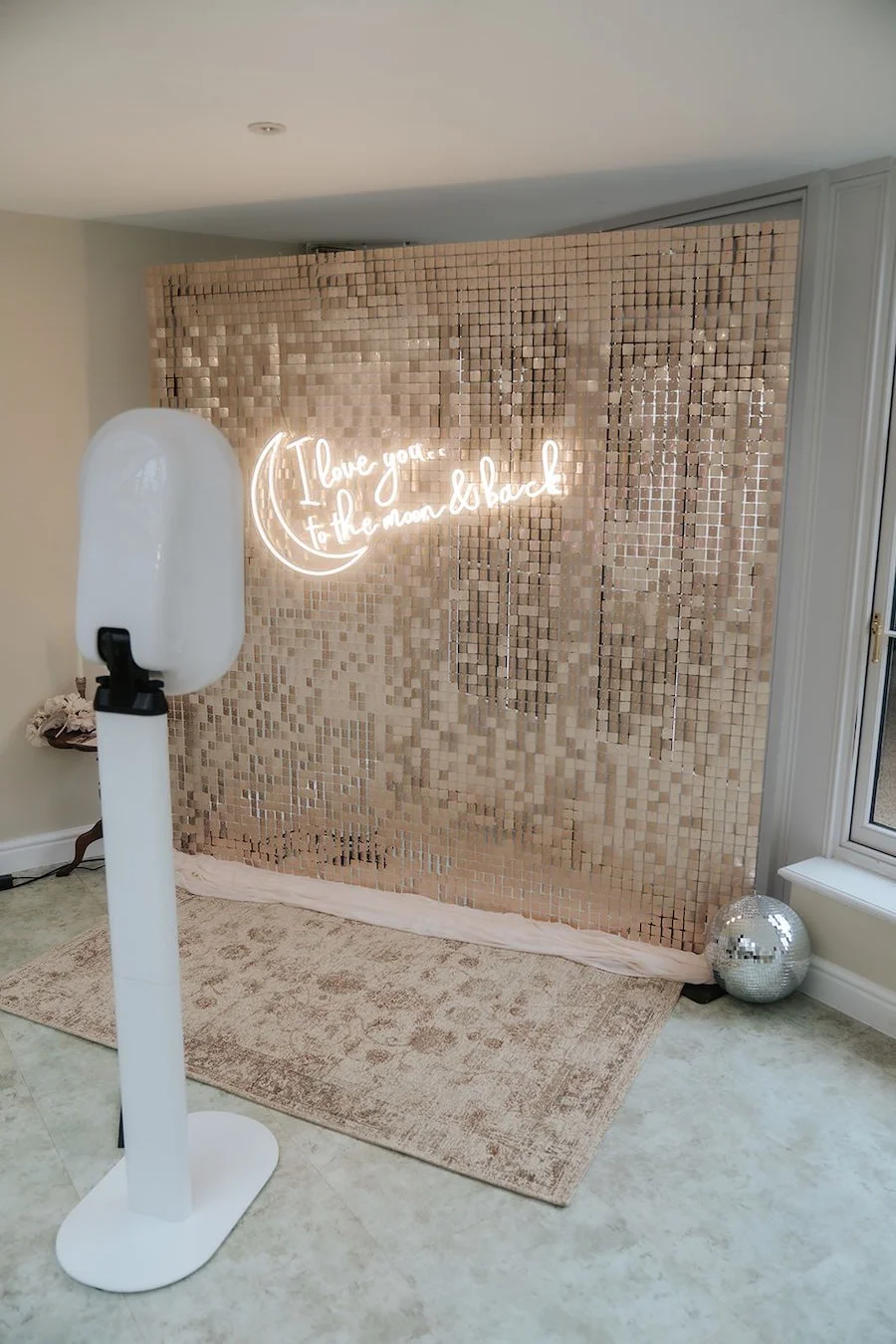 Decorative backdrop with a neon sign reading 'I Love You to the Moon and Back', small LEDs, beige mosaic tiles, a disco ball, a standing sanitizer or hand dryer, a pink rug, and a window in a room with neutral walls and tiled floor.