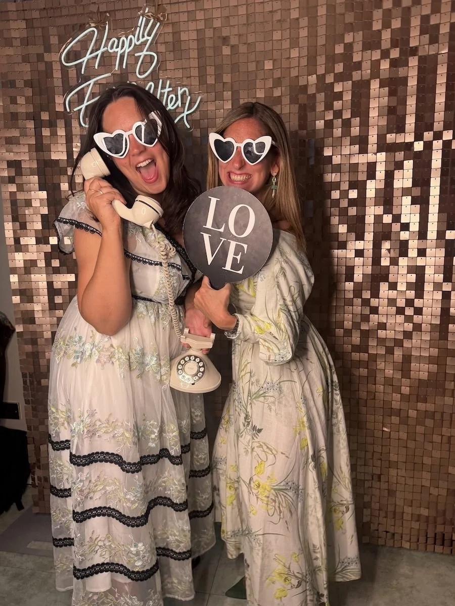Two women dressed in floral dresses wearing heart-shaped sunglasses, holding a vintage rotary phone and a round sign that says 'LOVE', standing in front of a wall with a shimmering, mosaic-like pattern and a sign that says 'Happily Ever After'.
