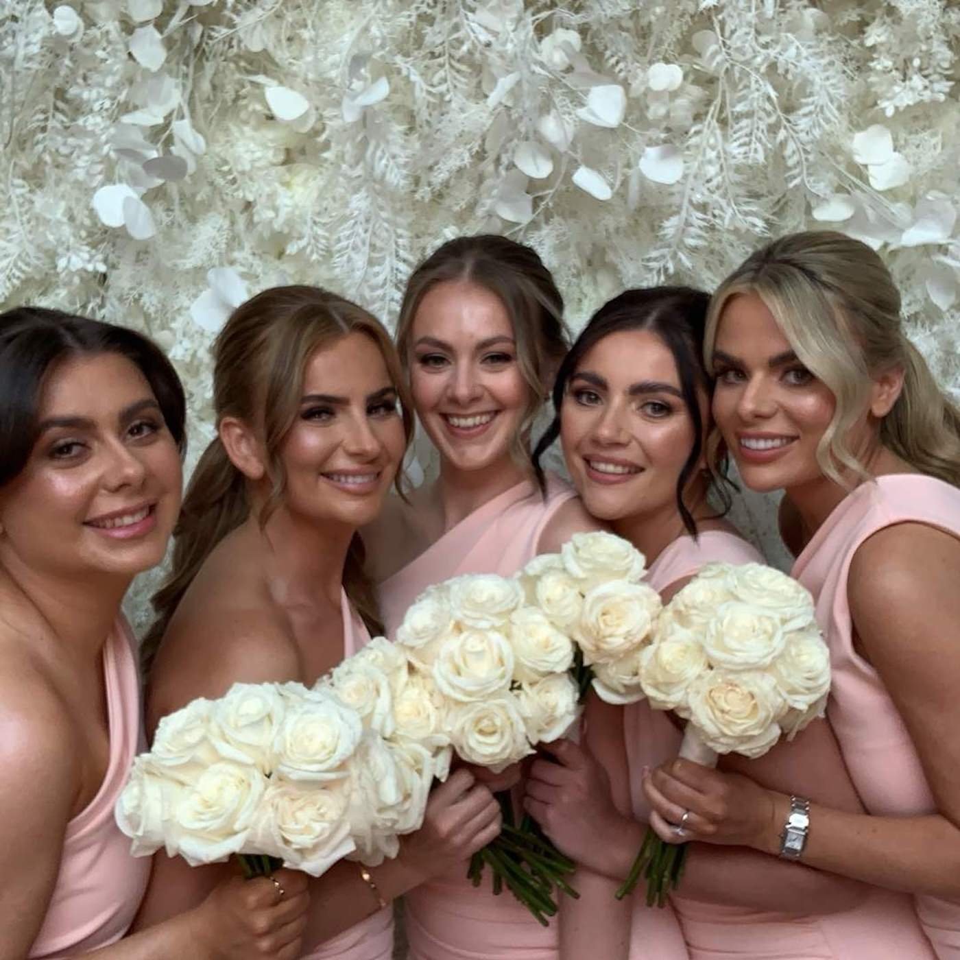 A group of six women wearing pink dresses, smiling, and holding white rose bouquets at a wedding celebration, standing in front of a decorative floral background.