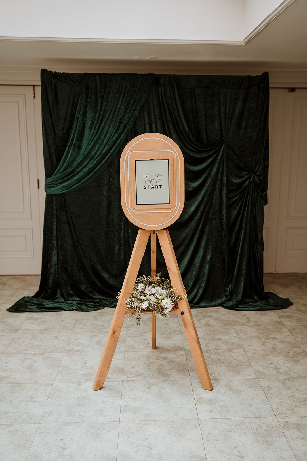 Wooden photo booth frame with daisies and greenery, green velvet curtain backdrop, sign on the frame that says 'tap to start'.