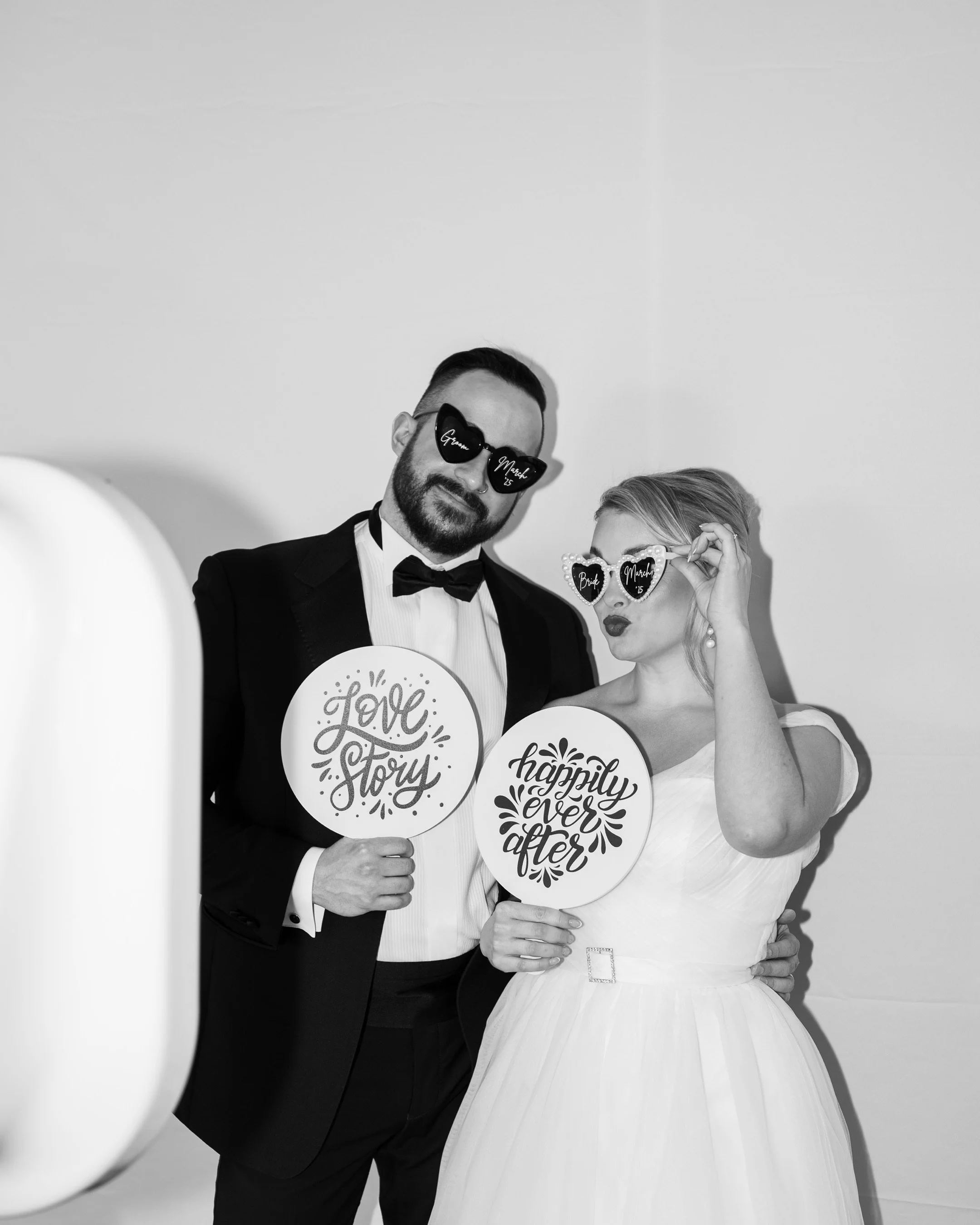 A wedding couple dressed in formal attire, the groom in a tuxedo and the bride in a wedding gown, holding circular signs with words like 'Love Story' and 'Happily Ever After.' They are wearing sunglasses with messages; the groom's say 'Groom' and the bride's say 'Bride,' and the bride is holding her sunglasses with her hand.