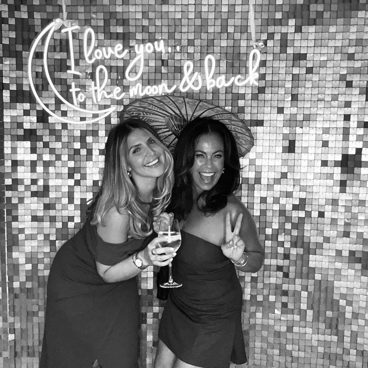 Two women in dresses posing in front of a mosaic wall with a neon sign that reads 'I love you to the moon and back'. One woman holds a drink, and the other makes a peace sign. They are smiling and enjoying a night out.
