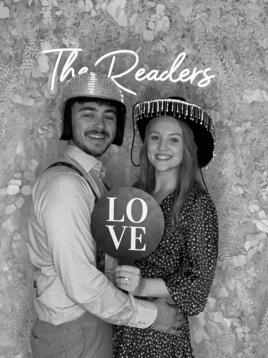 A couple is smiling at the camera while wearing hats with decorations and holding a balloon with the word 'LOVE'. They are standing in front of a floral backdrop with the words 'The Readers'.