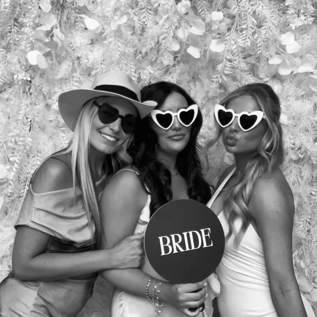 Three women at a wedding celebration, holding a sign that says 'BRIDE,' wearing sunglasses, standing in front of a decorative floral backdrop.