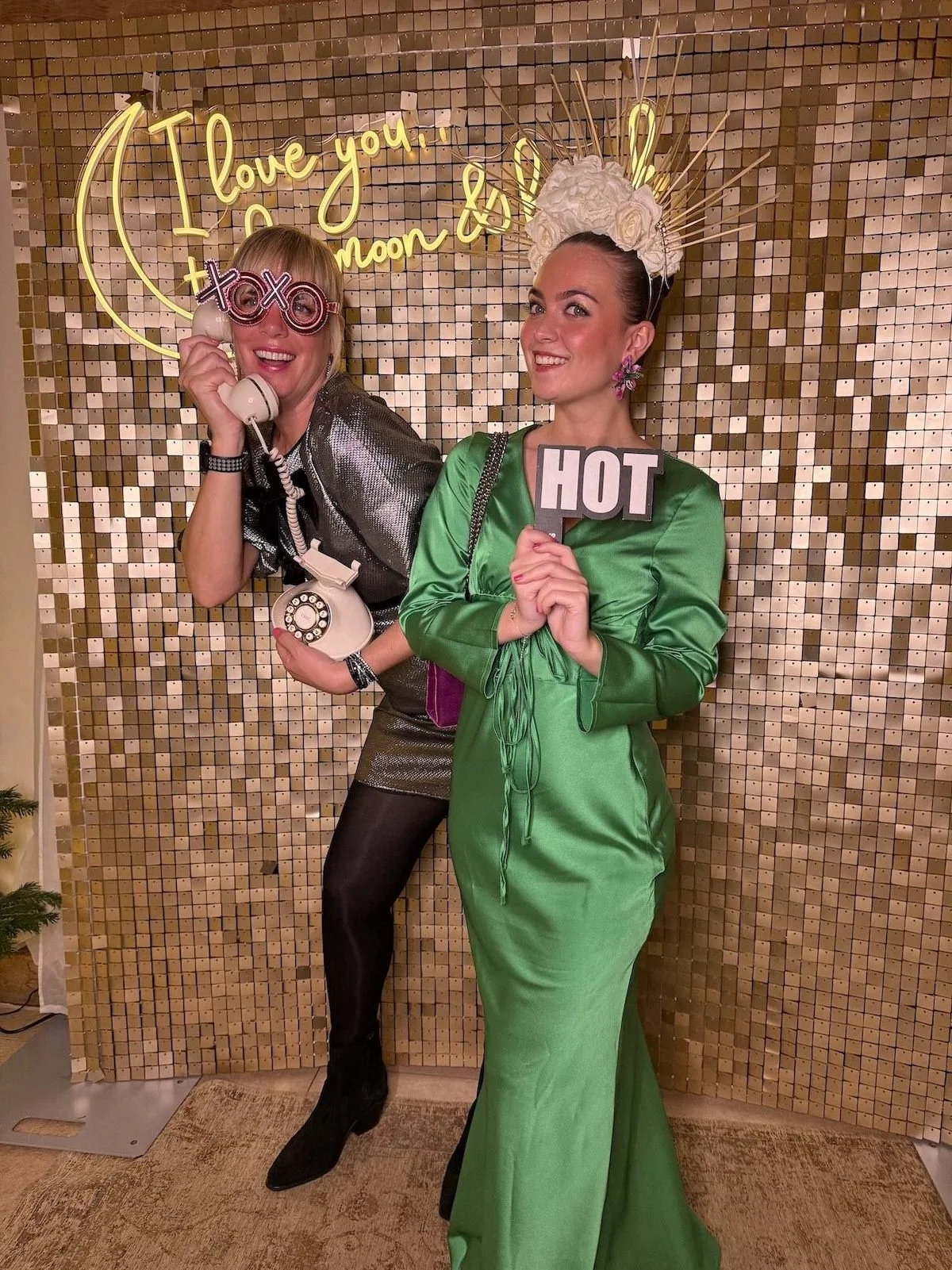 Two women posing for a photo against a gold mosaic wall with a neon sign that says 'I love you to moon & star.' The woman on the left is wearing a black and silver outfit with black tights, black boots, pink sunglasses that say 'toxo' and holding an 