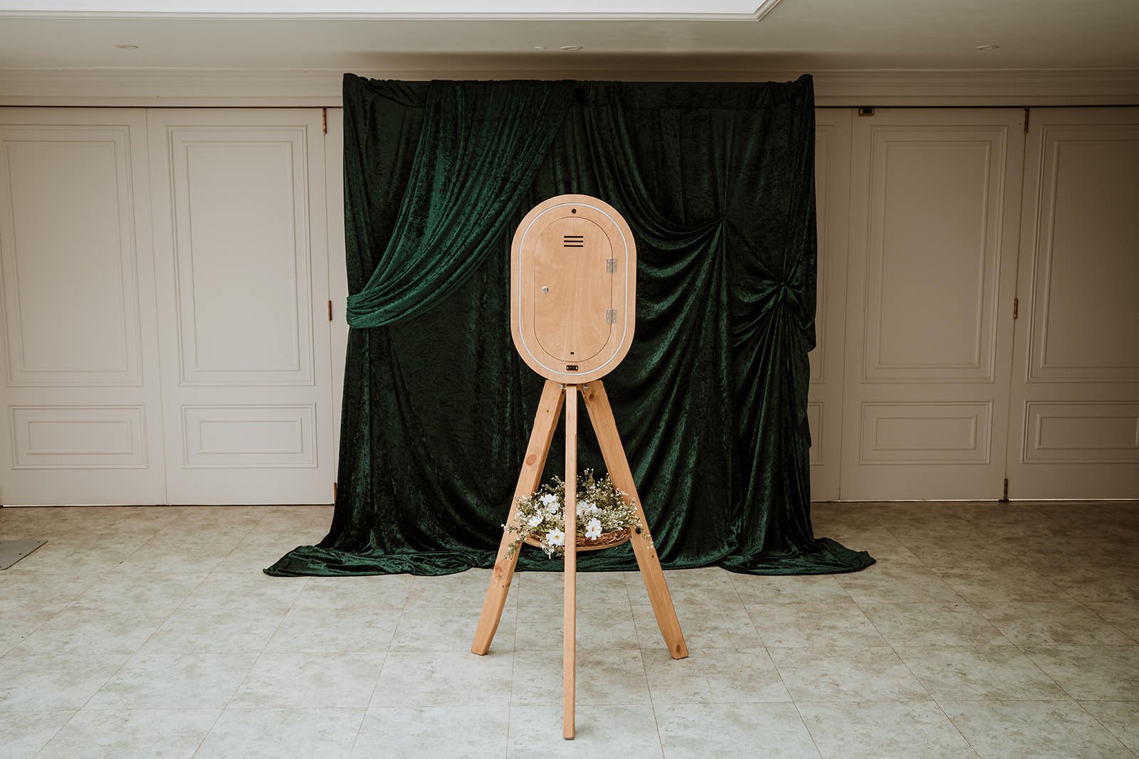 A wooden photo booth on tripod stand with a curtain backdrop and floral arrangement at the base of the stand.