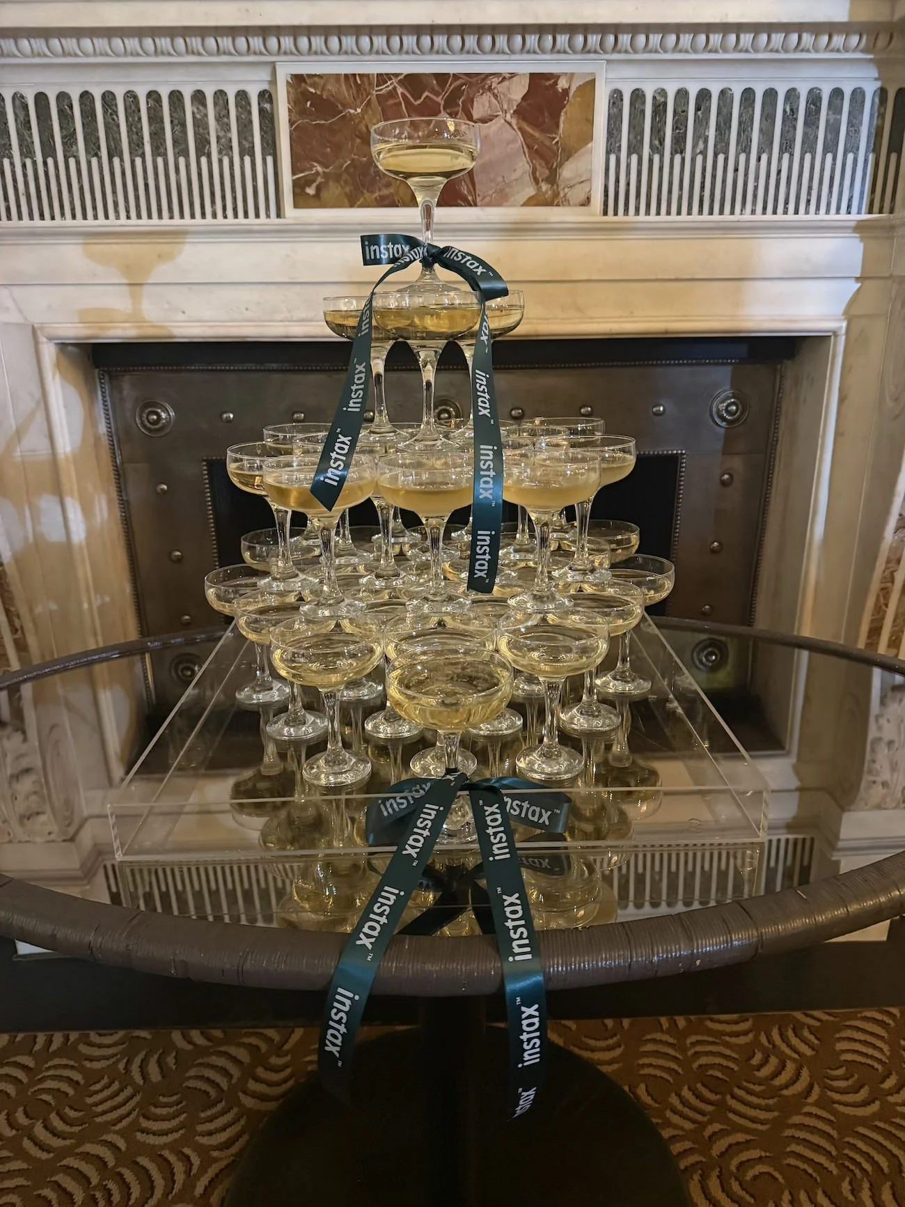 A pyramid of champagne glasses filled with champagne on a glass table, with a ribbon reading 'instax' draped over the top glass, in a room with ornate decor and a fireplace.