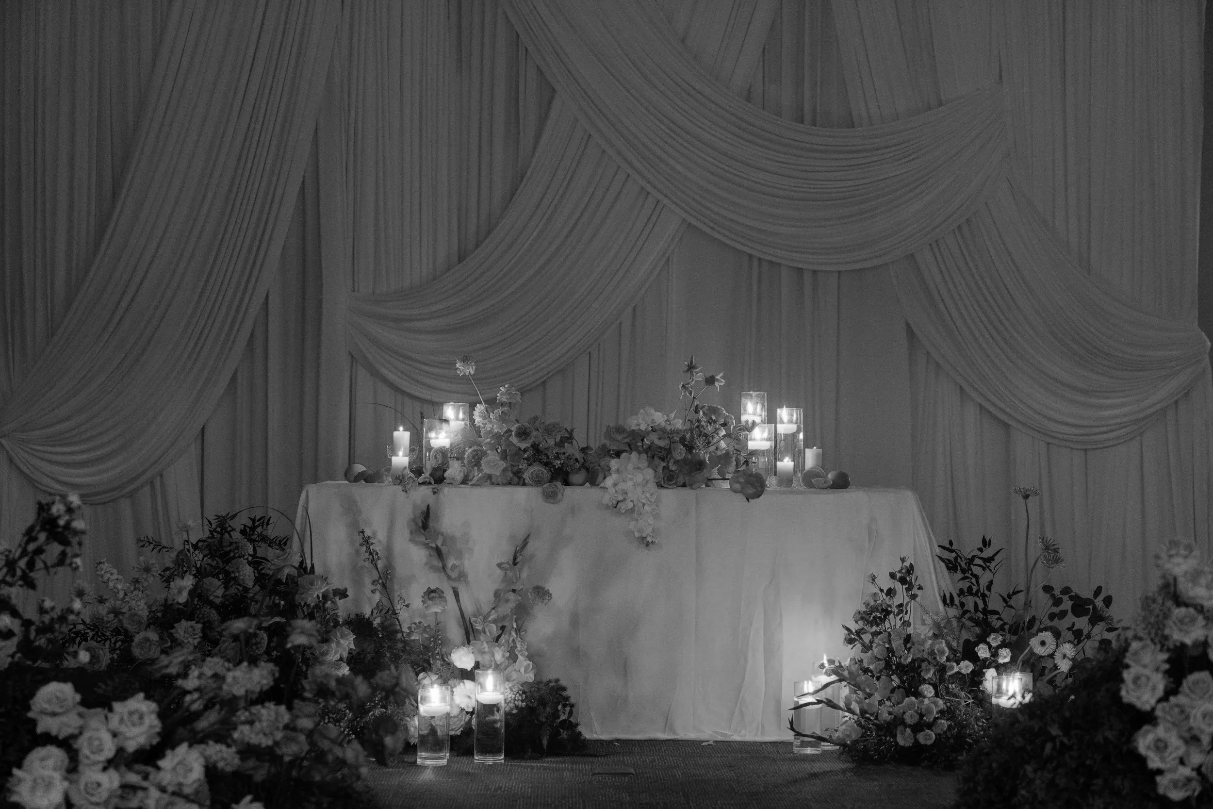 A decorated table with candles and floral arrangements in front of a backdrop with draped fabric, likely for a wedding or formal event.