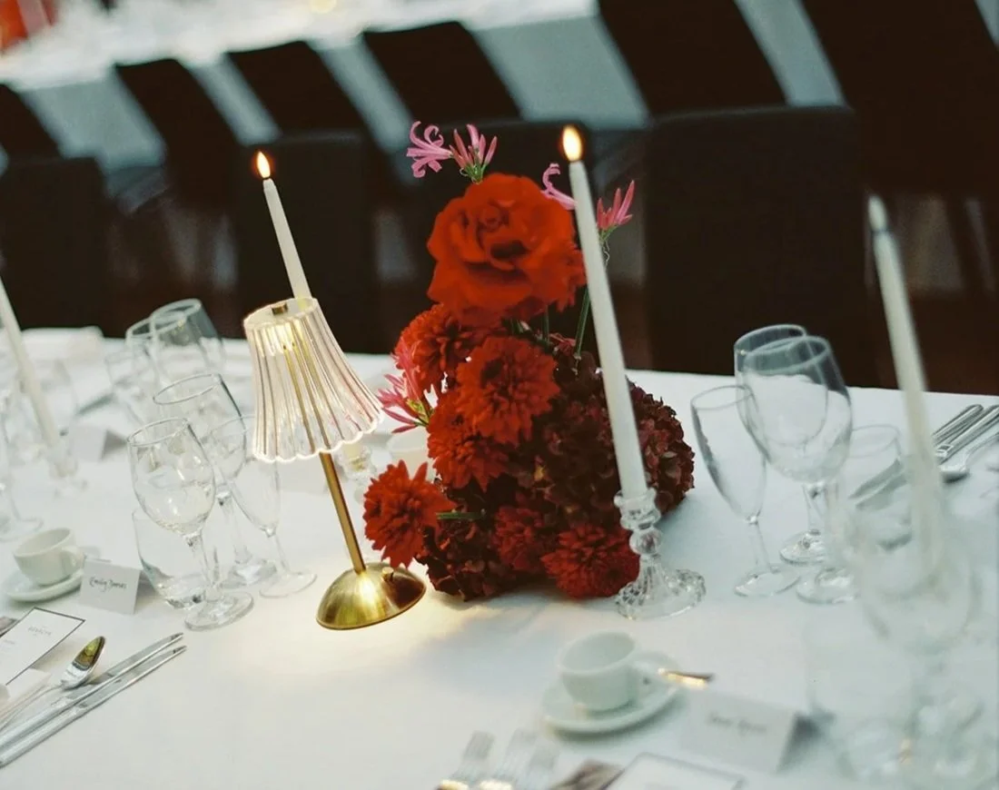 A close-up of an elegant dining table with a floral centerpiece, two lit candles, a small lampshade, and neatly arranged glassware and silverware.