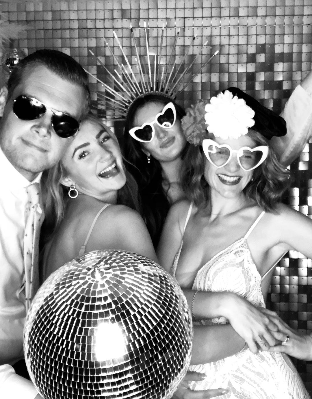 Group of four friends celebrating, wearing festive accessories, smiling, with a disco ball in the foreground and a sparkly wall background.