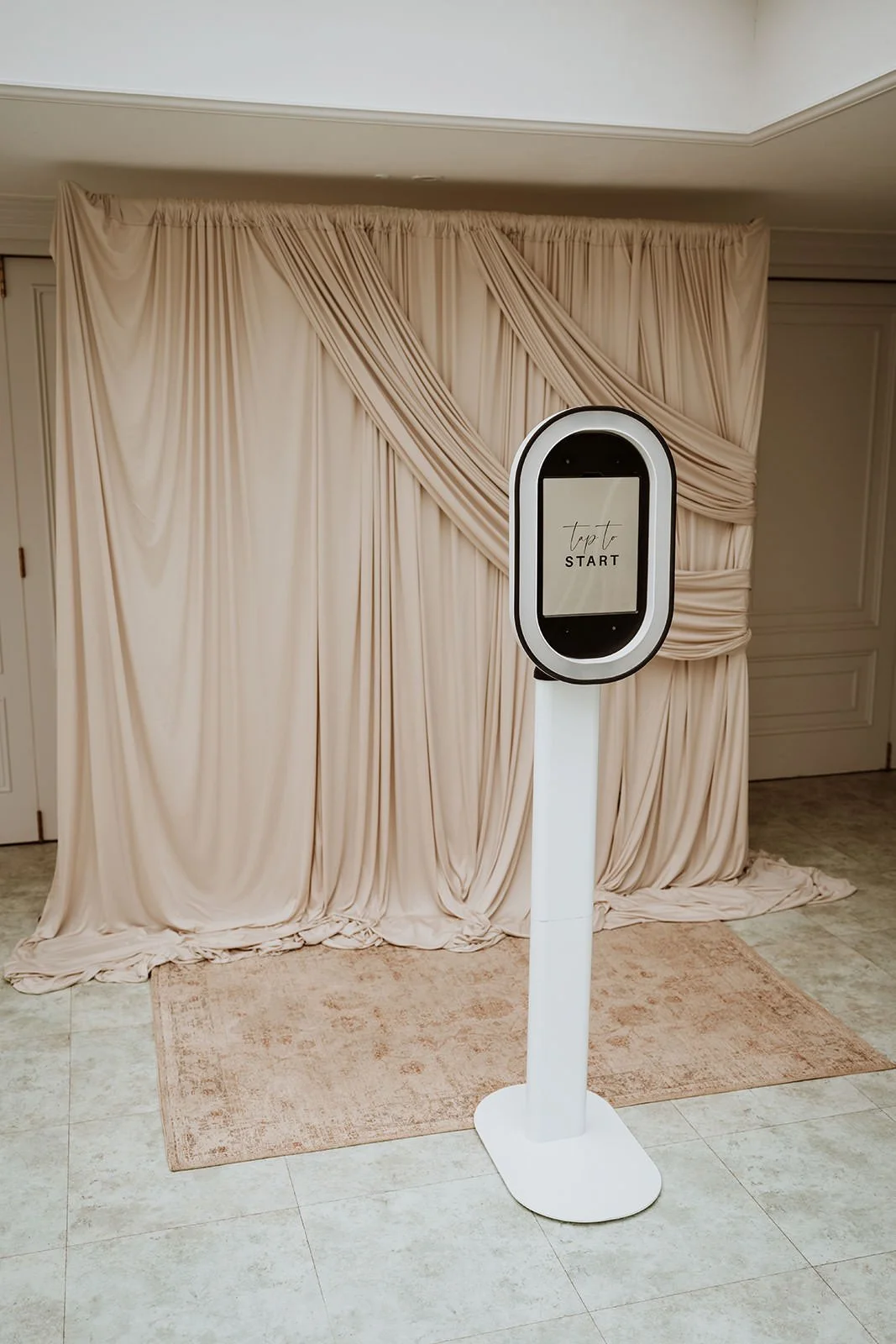 An electronic photo booth with a screen displaying 'Tap to start' stands in front of cream-colored drapery arranged as a backdrop, on a beige rug.